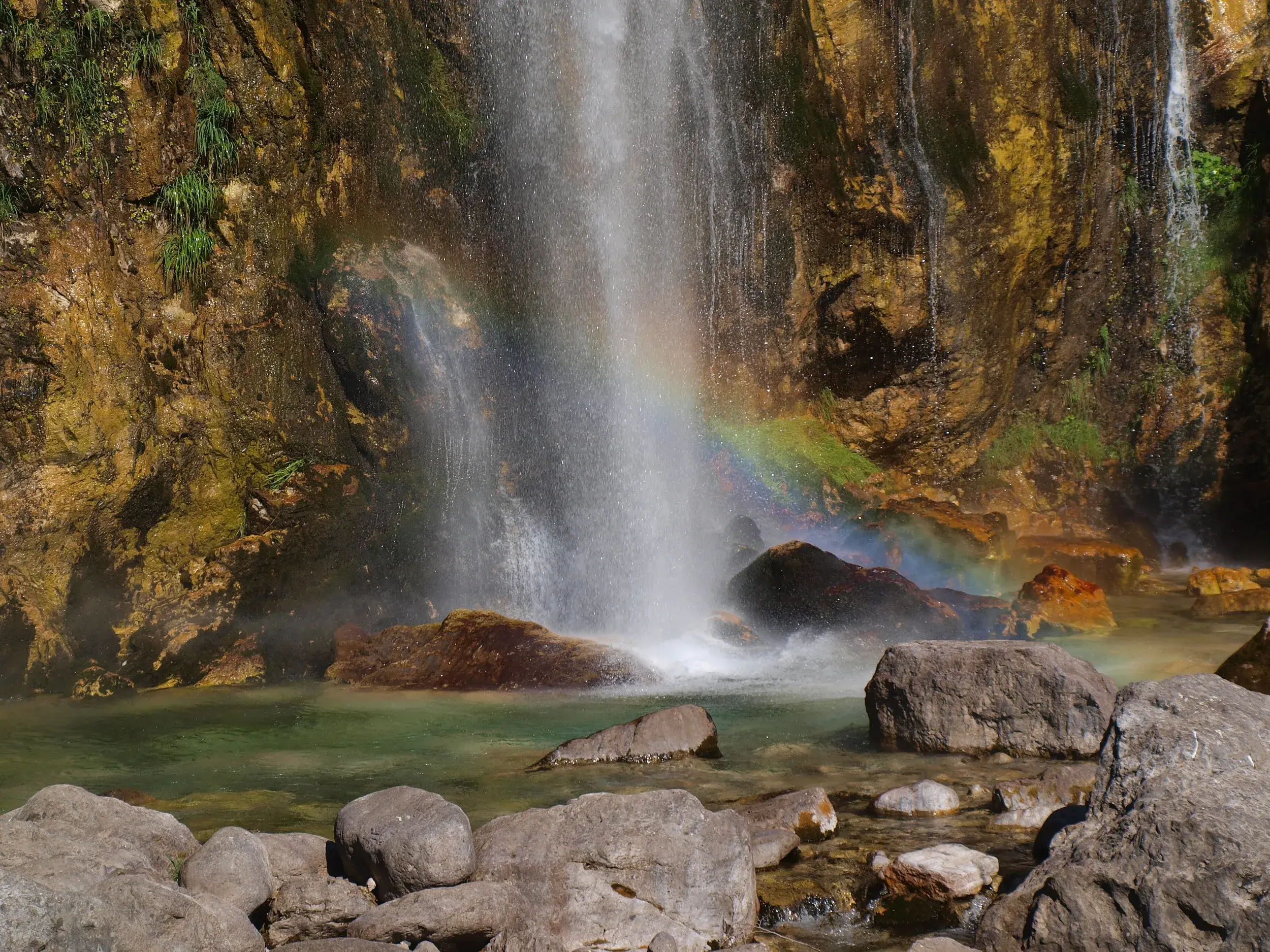 Regenbogen in der Gischt des Grunas-Wasserfalls, der über eine Felswand in ein türkises Becken in den albanischen Alpen stürzt.