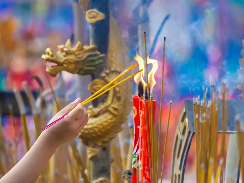 Hand entzündet gelbe Räucherstäbchen an einer Flamme in einem Tempel in China, umgeben von Rauch und goldenen Drachenfiguren.