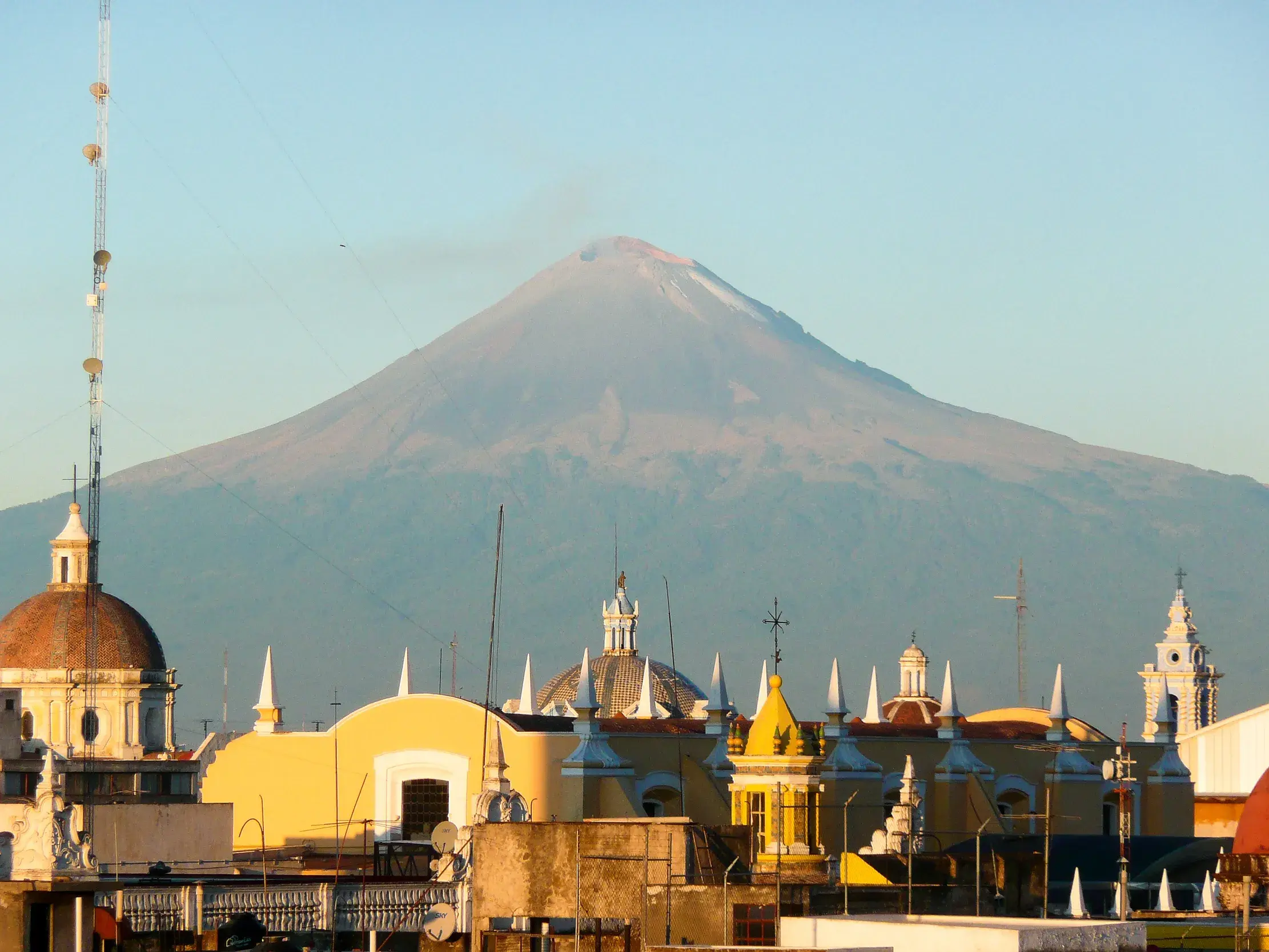 Gelbe Kirchtürme und Dächer von Puebla vor dem Gipfel des leicht rauchenden Vulkans Popocatépetl in Mexiko.