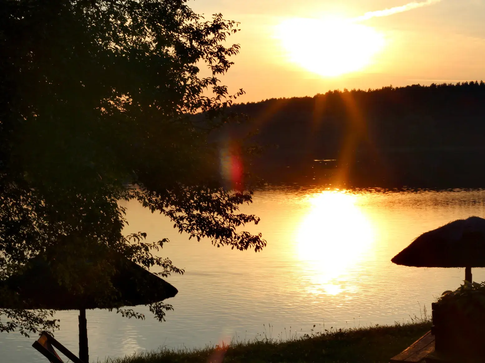 Stimmungsvolle Abenddämmerung am Czos-See in Masuren. Die untergehende Sonne spiegelt sich im Wasser vor einem bewaldeten Ufer.