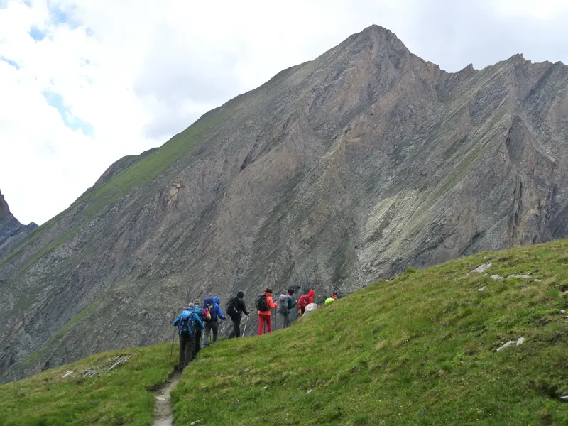Eine Trekking-Gruppe steigt auf einem schmalen Pfad über eine grüne Wiese durch die Berglandschaft des Nationalparks Hohe Tauern.