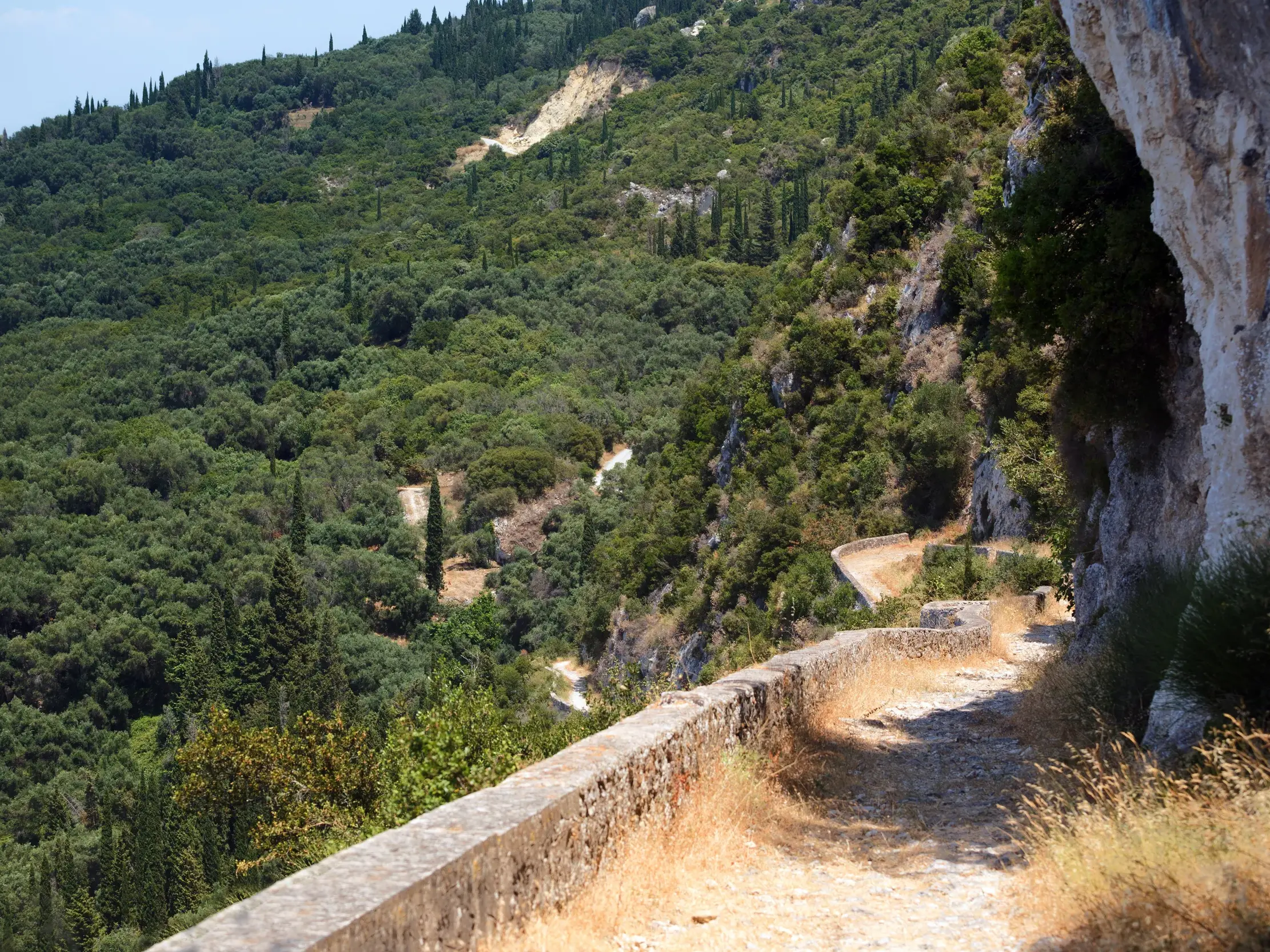 Auf dem Korfu-Trail führt ein historischer Pfad mit Steinmauer durch die bergige, grüne Landschaft mit Zypressen.