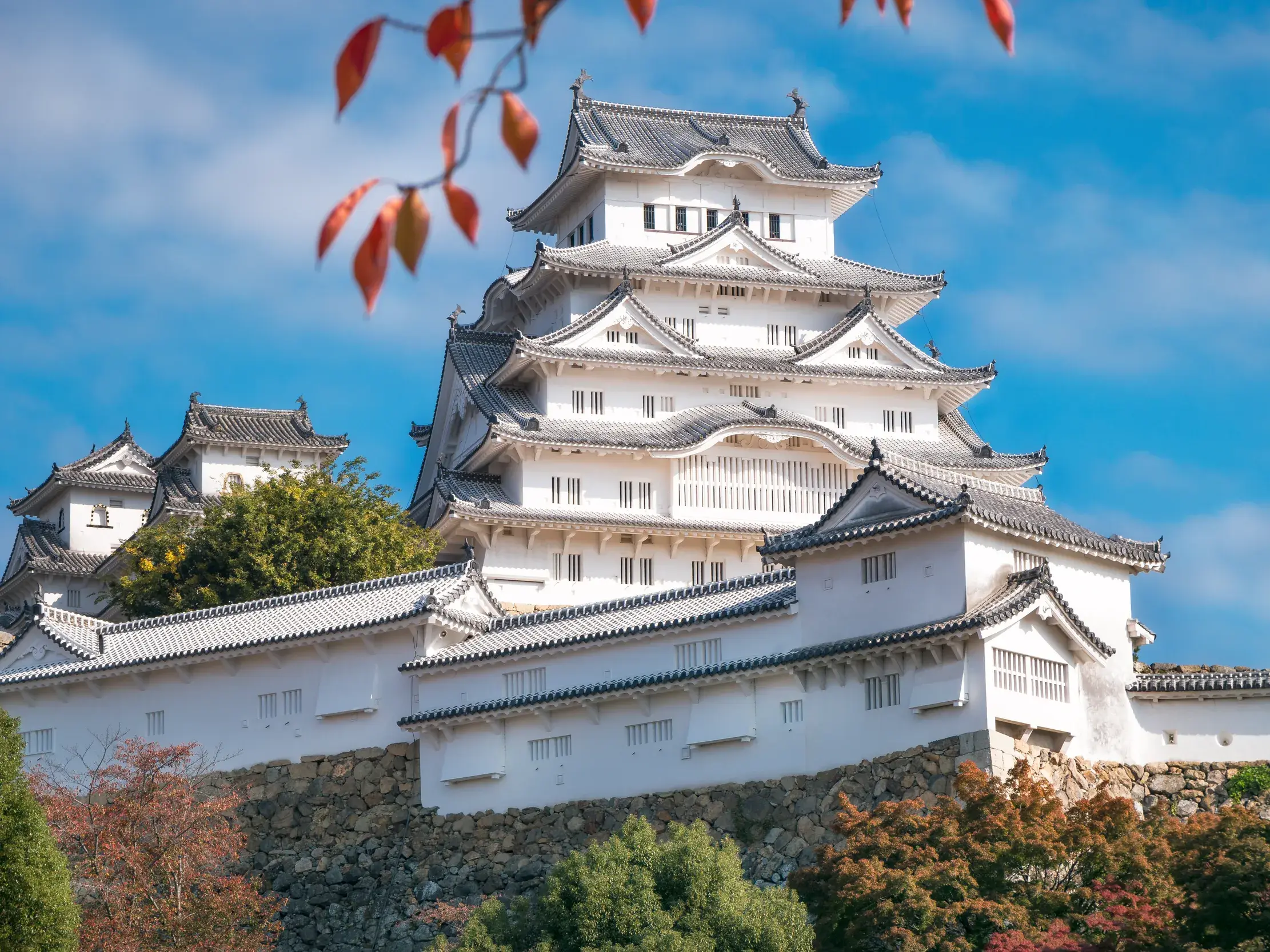 Die majestätische, weiße Burg des Weißen Reihers in Himeji, umrahmt von rotem Herbstlaub unter strahlend blauem Himmel.