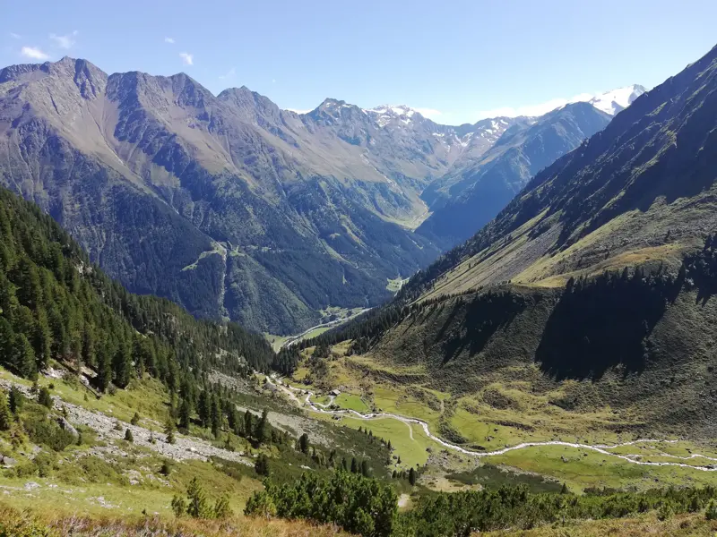 Weitblick beim Trekking auf der Alpenüberquerung in ein sonniges Alpental mit Fluss, umrahmt von den Stubaier Alpen.