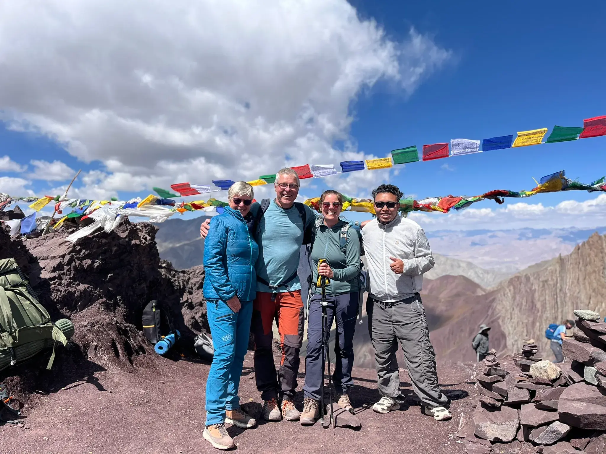 Glückliche Hauser-Wandergruppe mit Reiseleiter posiert auf dem Gipfel des Stok La in Ladakh unter bunten Gebetsfahnen.