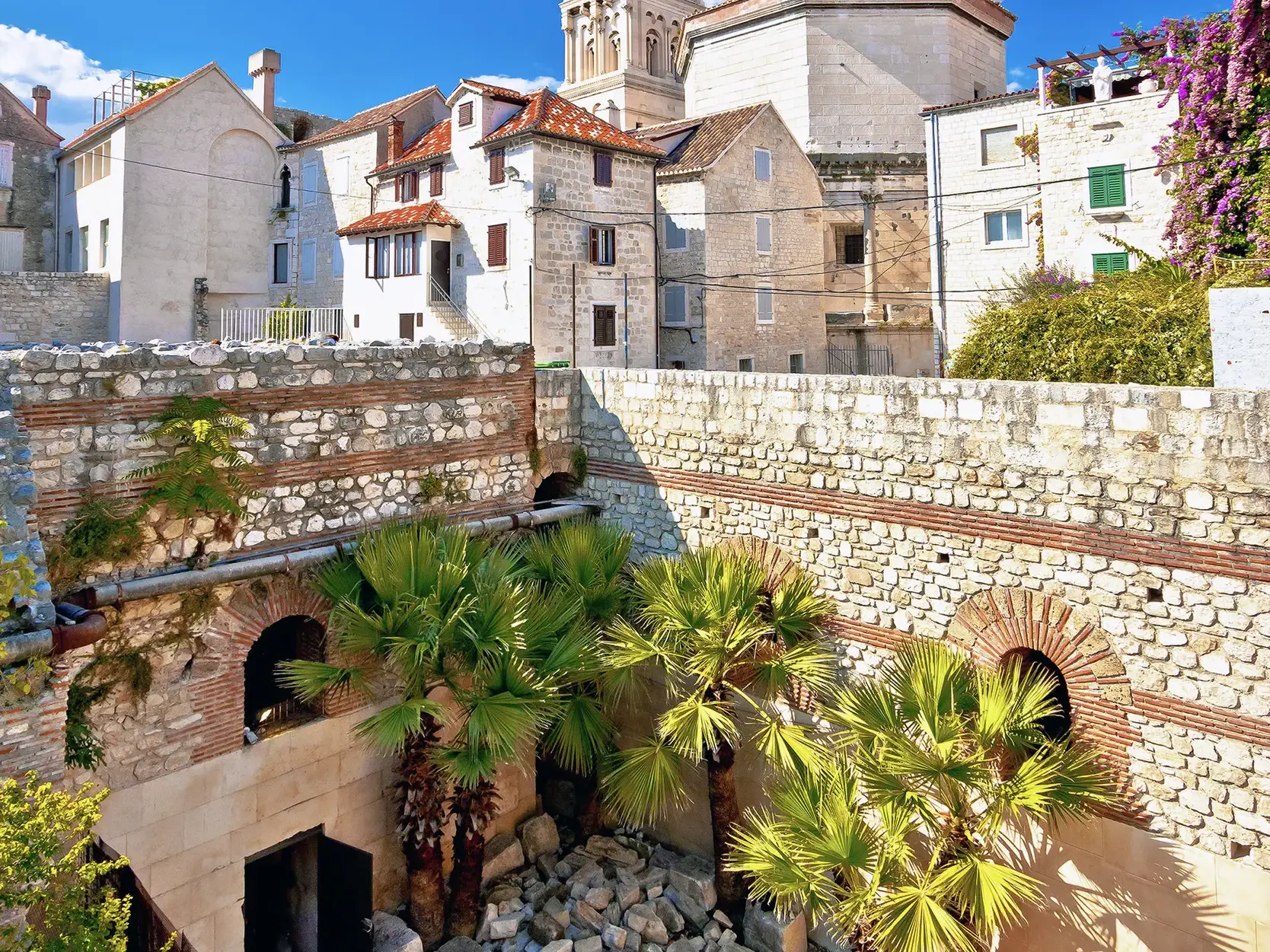 Blick über die steinernen Ruinen und Palmen im Diokletian-Palast auf die historischen Gebäude der Altstadt von Split.