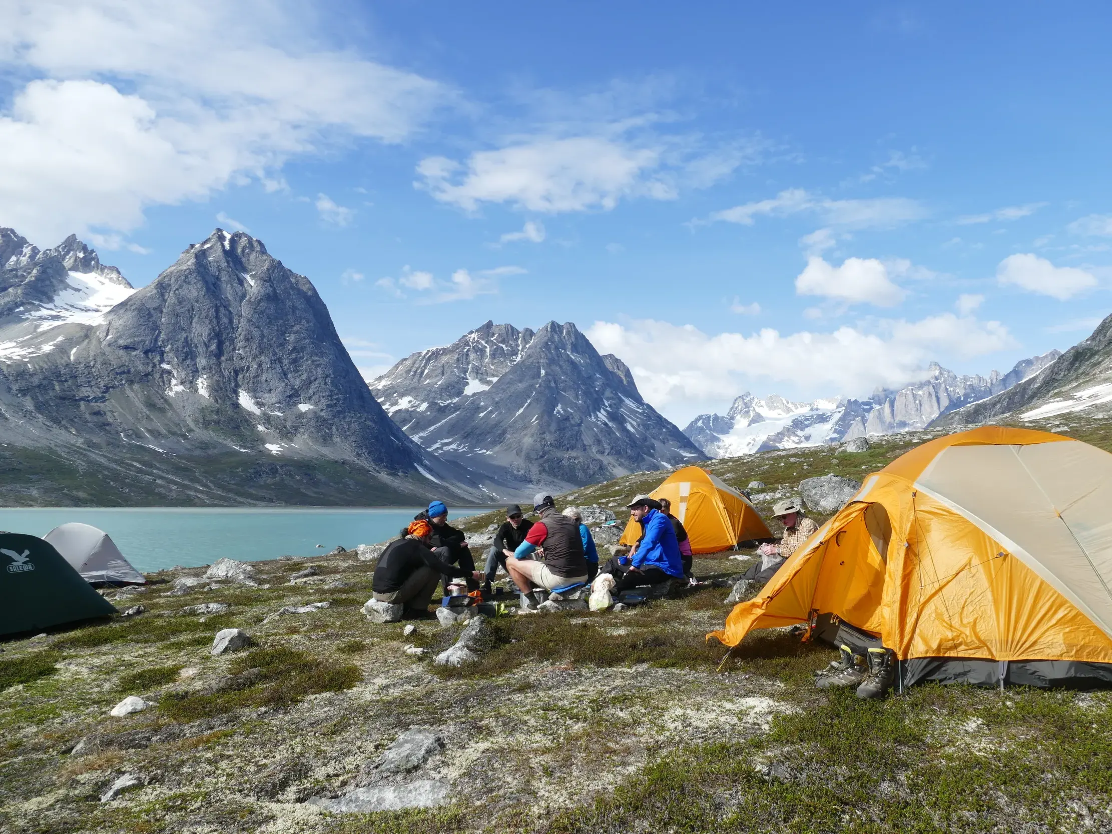 Eine Trekkinggruppe rastet in ihrem Zeltlager am Ufer eines Fjords in der wilden Berglandschaft Ostgrönlands.