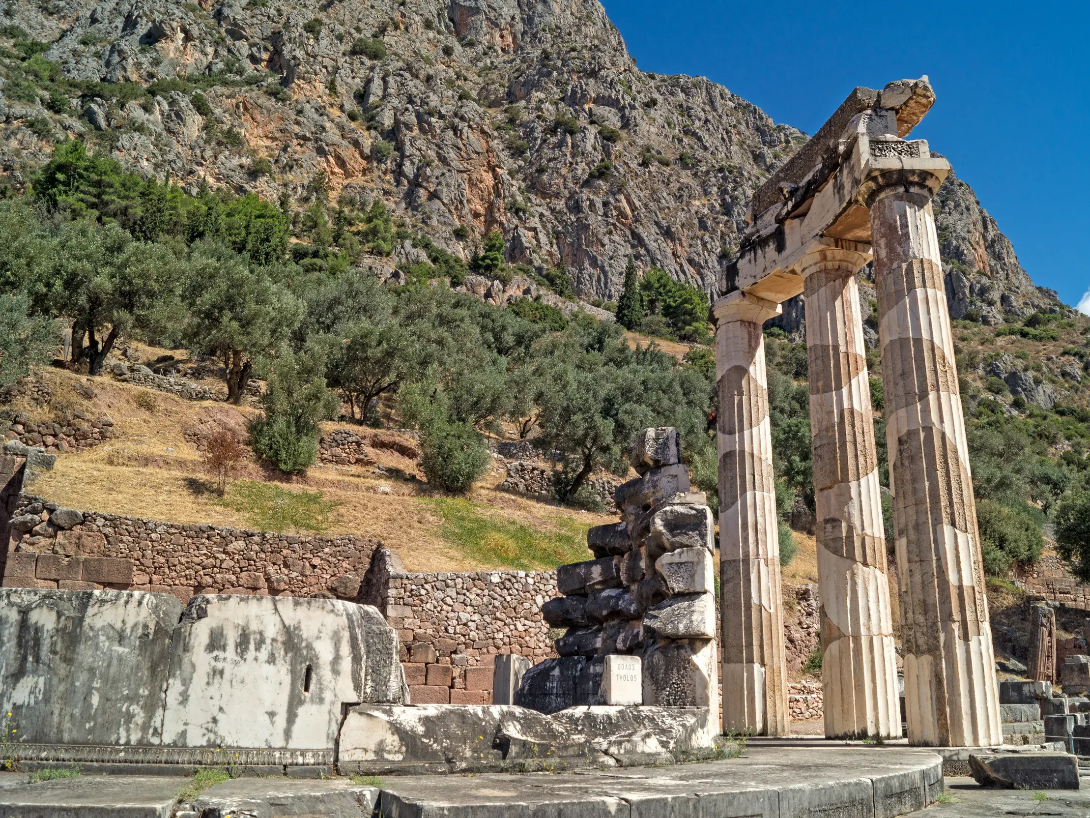 Die Marmorsäulen des Tholos von Delphi, UNESCO-Welterbe, vor der felsigen Kulisse des Parnassgebirges an einem sonnigen Tag.