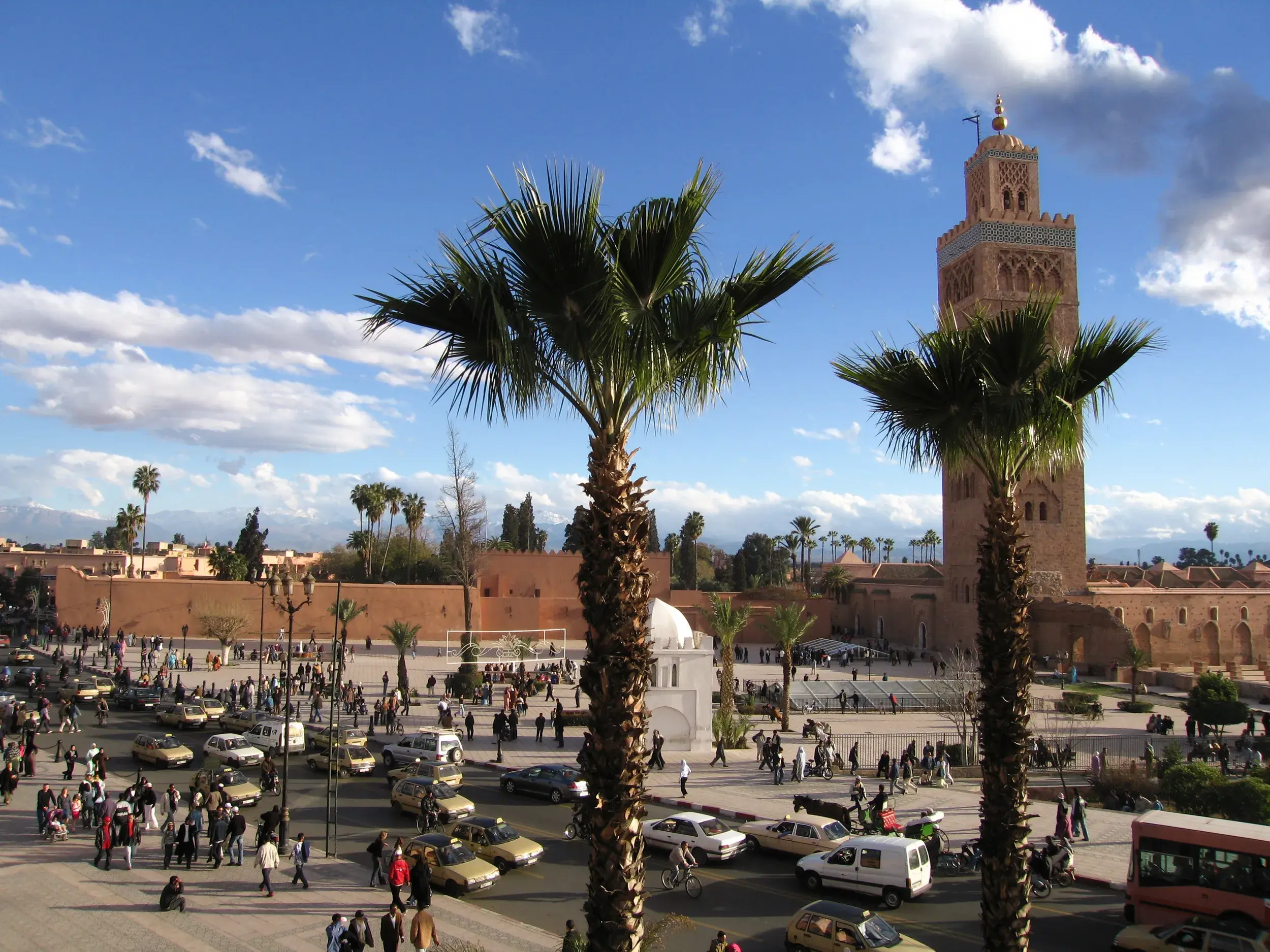 Belebter Djemaa el Fna Platz in Marrakesch mit der Koutoubia-Moschee und dem fernen Atlasgebirge im Hintergrund.