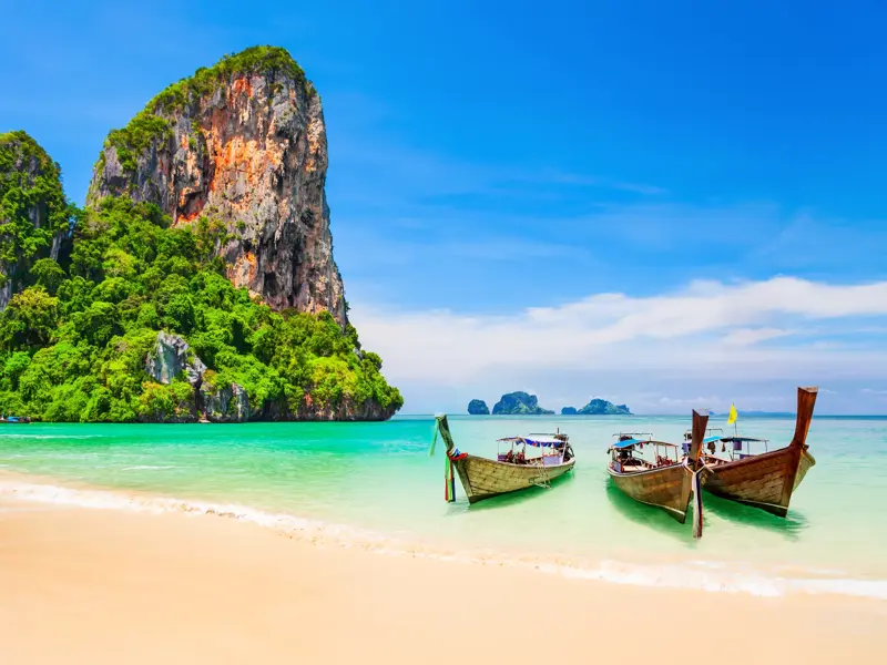 Drei hölzerne Langschwanzboote im seichten, türkisfarbenen Wasser am Sandstrand von Khao Lak vor einer hohen, bewachsenen Felsklippe.