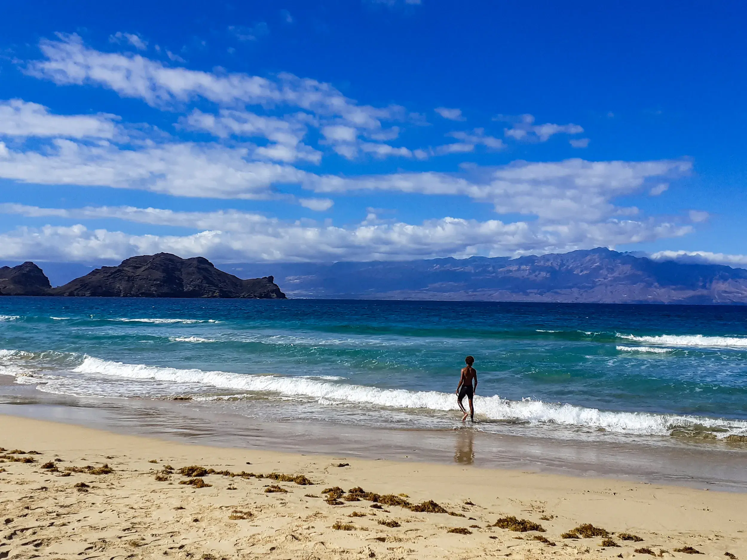 Eine Person watet am Sandstrand von São Vicente ins türkise Meer, im Hintergrund die beeindruckende Berglandschaft der Kapverden.