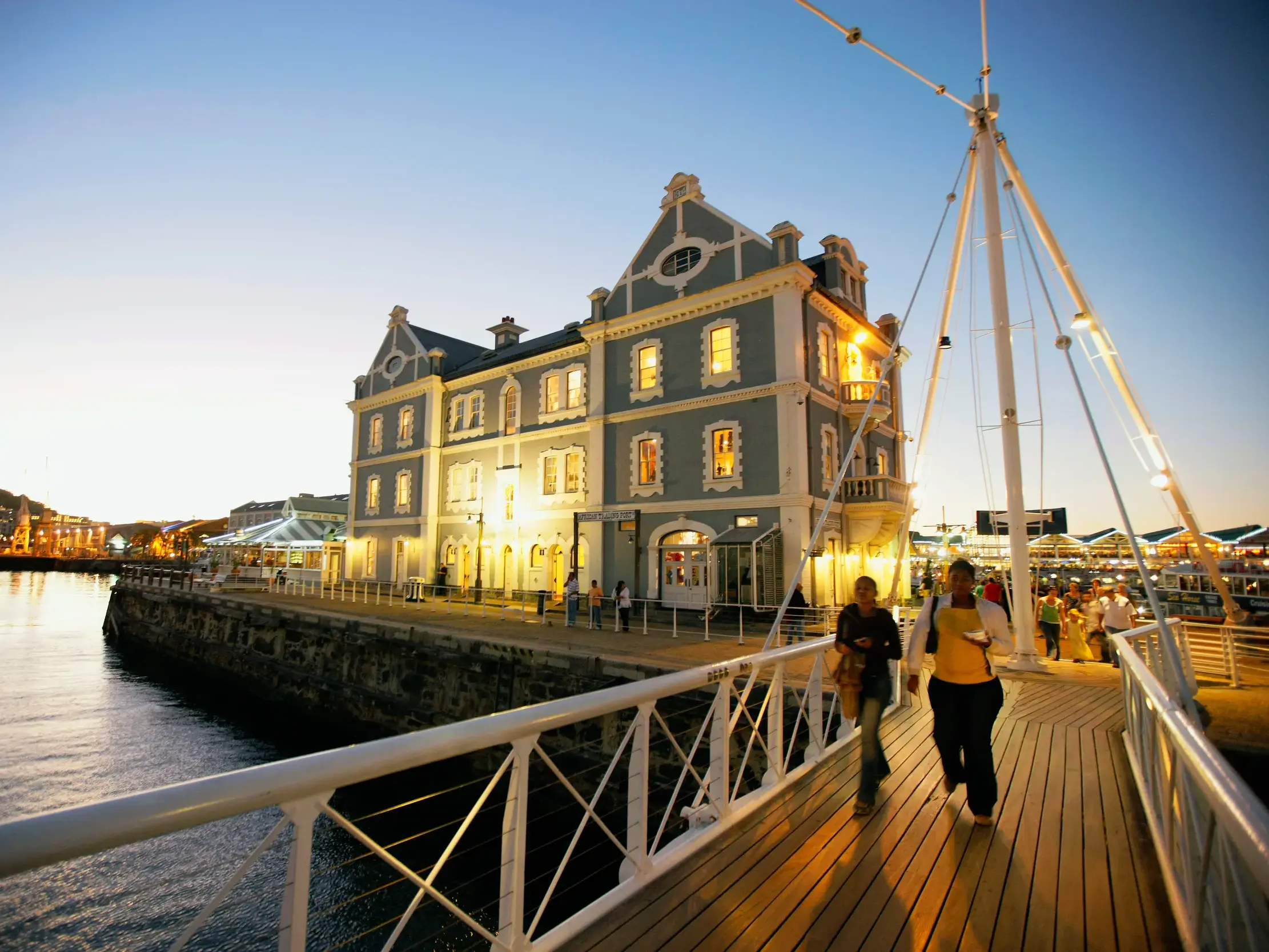 Illuminiertes historisches Gebäude an der V&A Waterfront in Kapstadt mit Besuchern auf einer Brücke in der Abenddämmerung.