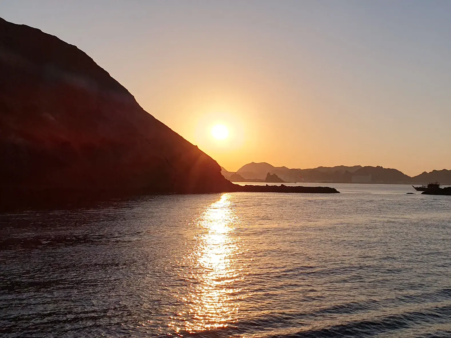 Die Sonne versinkt am Horizont und taucht die Küste bei Maskat während einer Dhaufahrt in warmes, goldenes Licht.