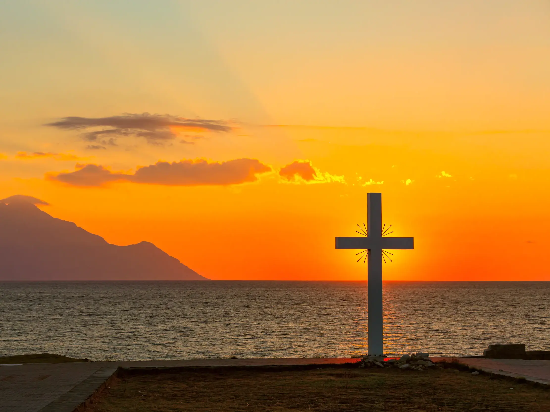 Stimmungsvoller Sonnenuntergang am Meer mit weißem Kreuz an der Küste und der Silhouette des Berges Athos am Horizont.