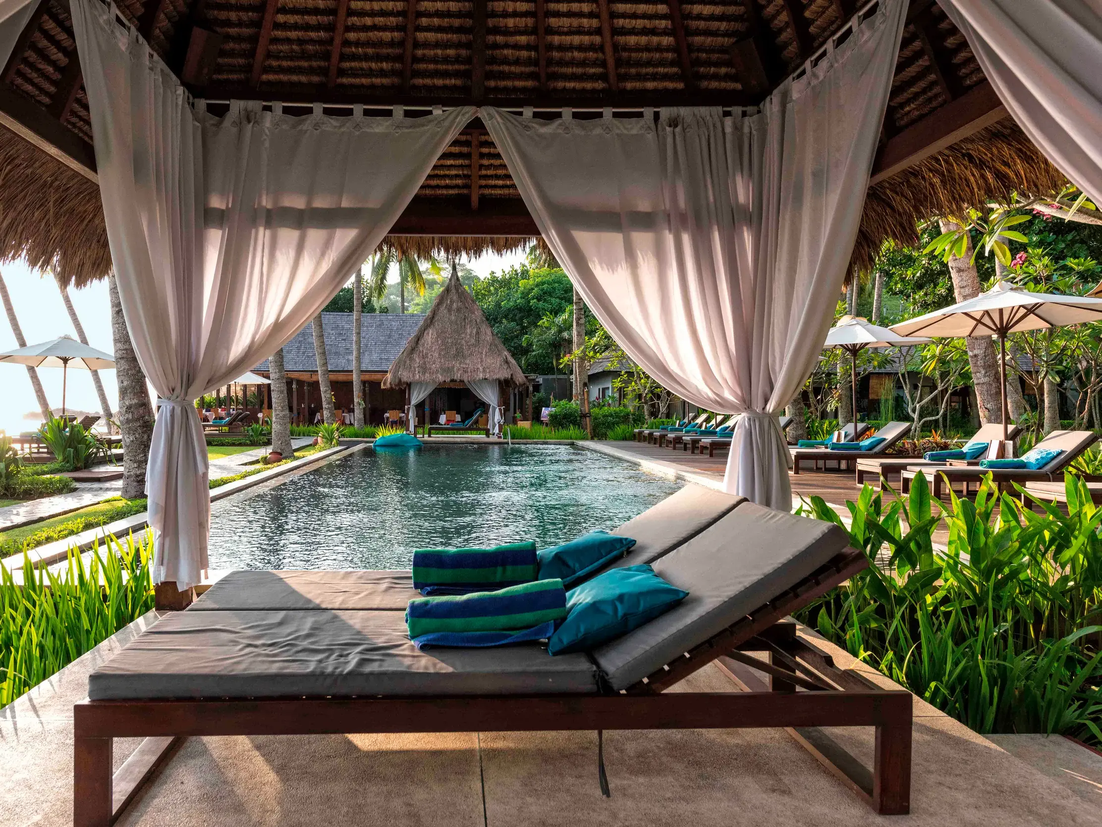 Blick aus einer Cabana mit Sonnenliegen auf den Pool des Boutique-Hotels Chandi auf Lombok bei Sonnenuntergang.