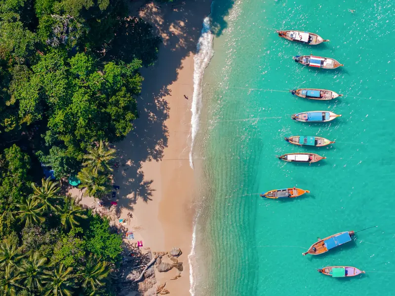 Vogelperspektive auf einen Traumstrand in Phuket mit Palmen und Longtail-Booten, die im türkisfarbenen Meer ankern.