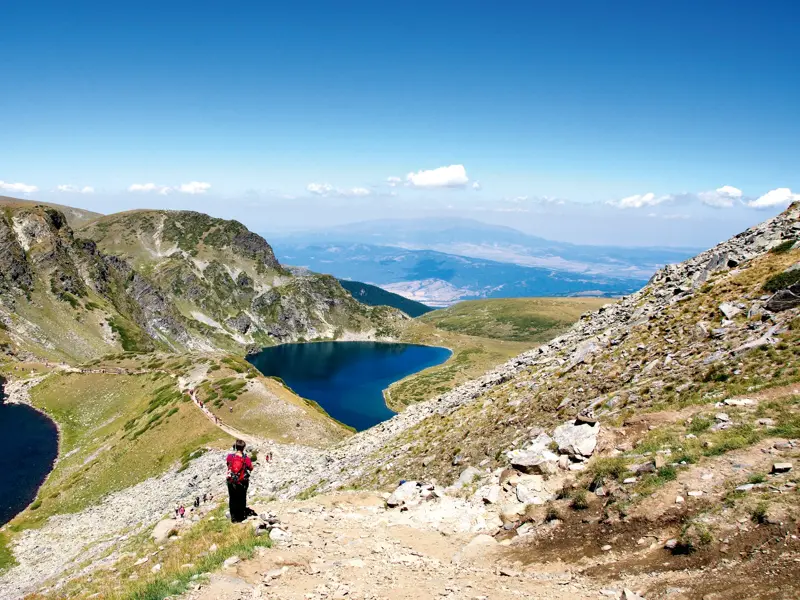Wanderer auf einem Bergpfad genießen den Panoramablick auf die tiefblauen Sieben Rila-Seen im Rila-Gebirge Bulgariens.