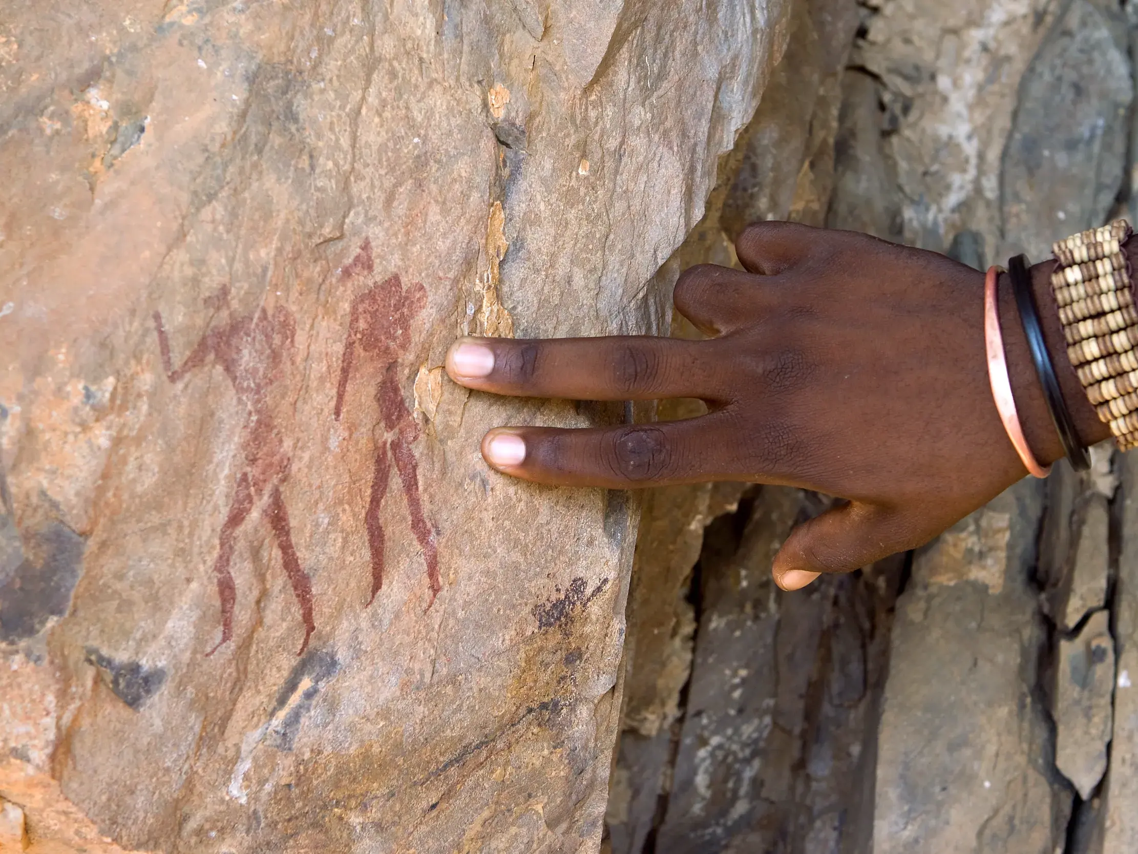 Nahaufnahme einer Hand mit Armbändern, die auf die prähistorischen Felsmalereien in Twyfelfontein, Namibia, zeigt.