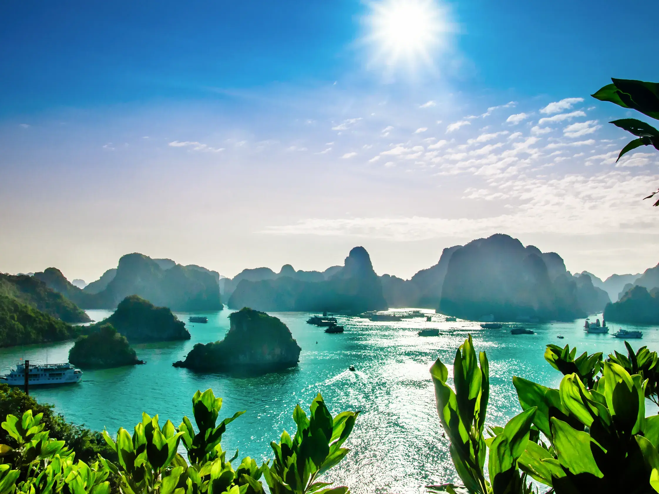 Strahlender Sonnenschein über der türkisen Halongbucht in Vietnam mit Ausflugsbooten, die zwischen den berühmten Karstfelsen fahren.