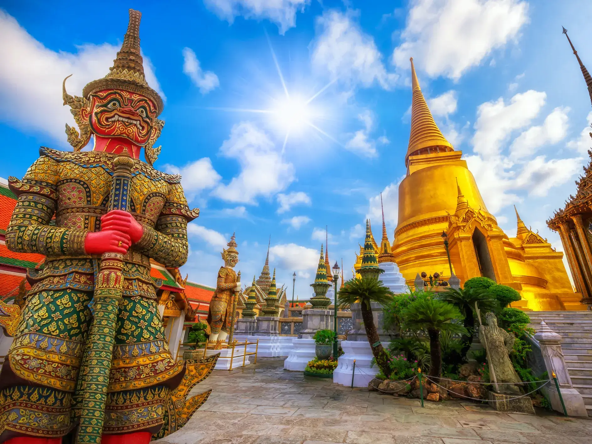Ein roter Tempelwächter bewacht den Königspalast in Bangkok mit seinen goldenen Pagoden bei strahlendem Sonnenschein.