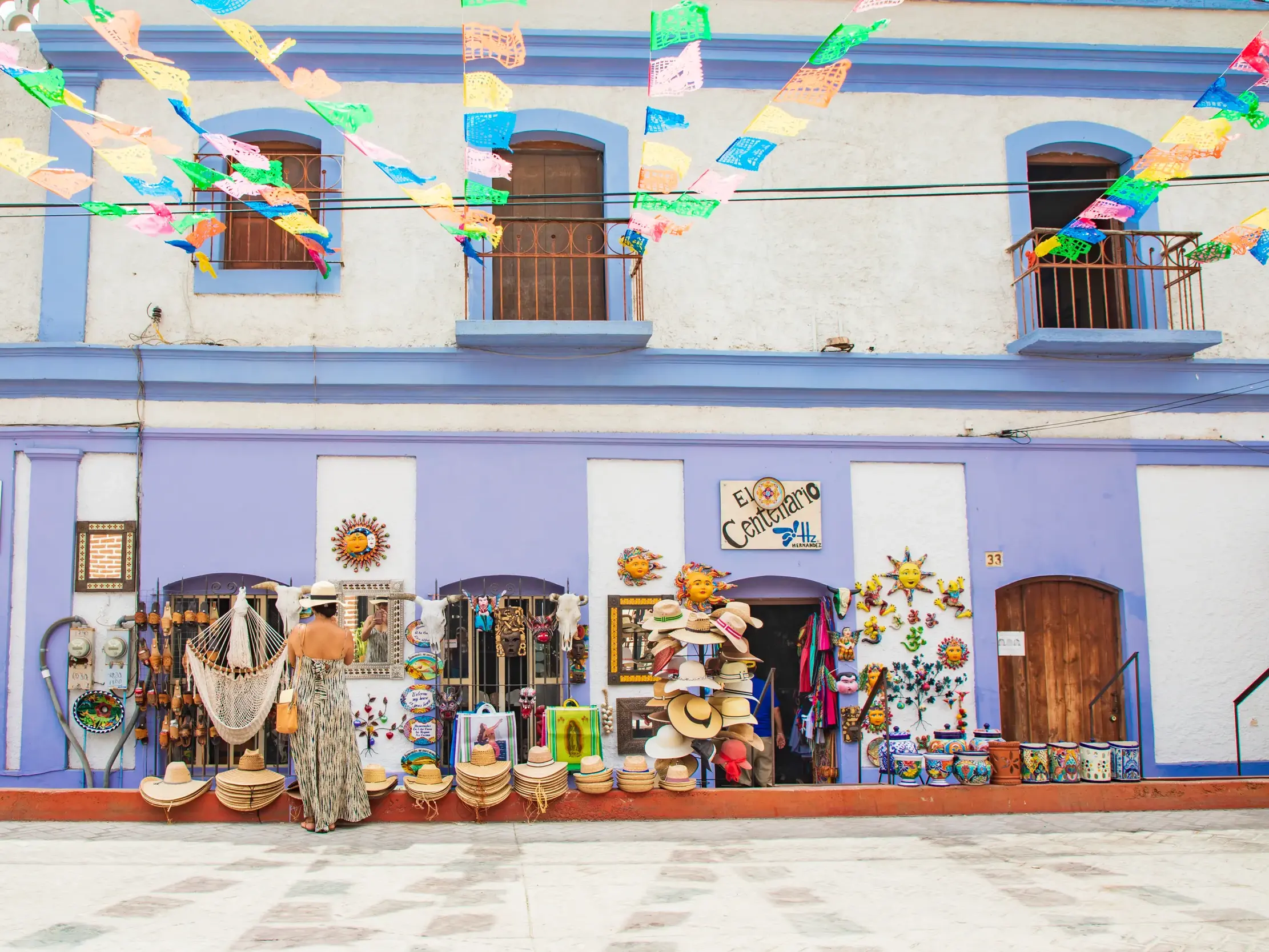 Eine Frau mit Sommerhut betrachtet lokales Kunsthandwerk vor einem bunten Kolonialgebäude in San José del Cabo, Mexiko.