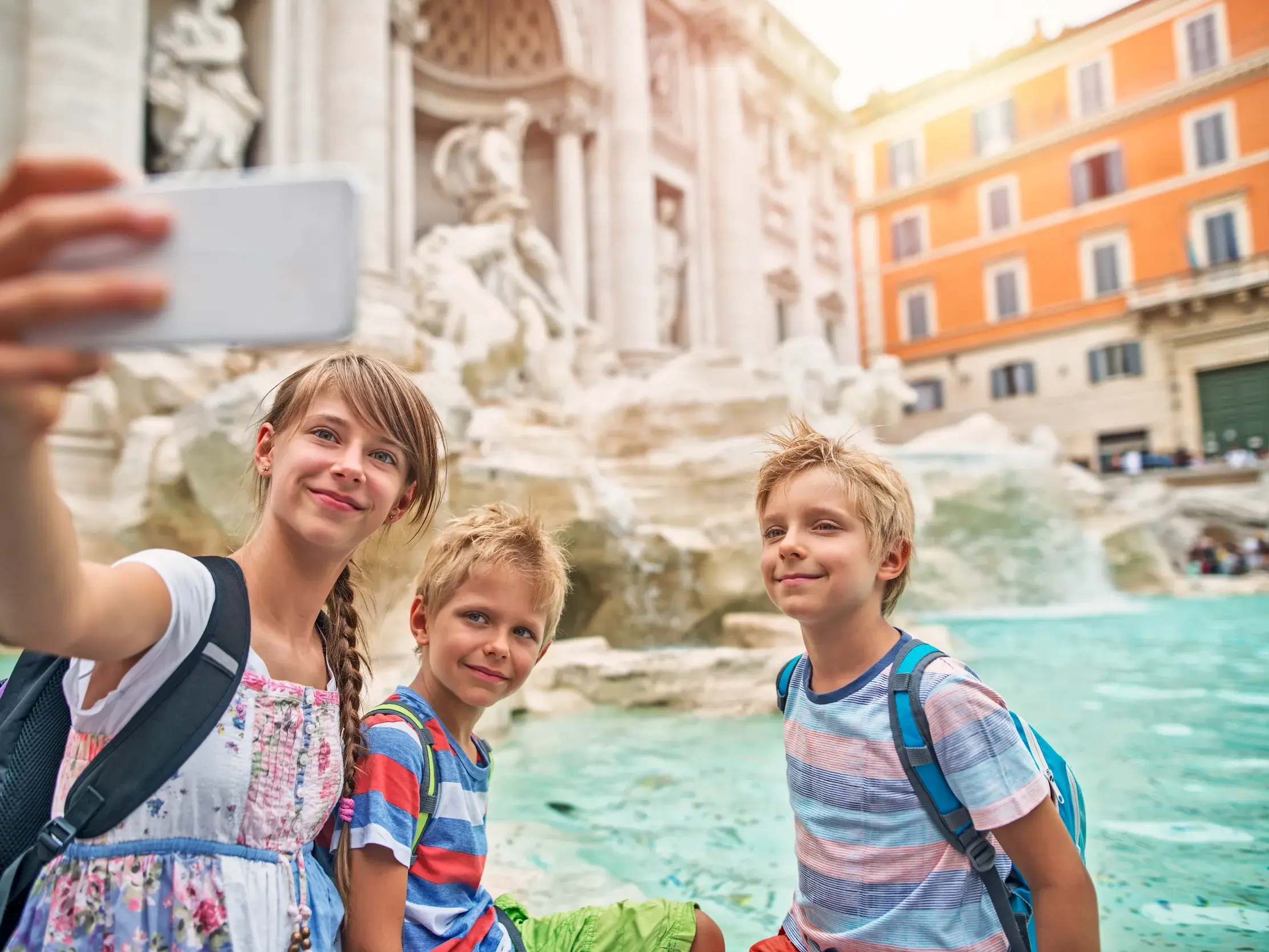 Drei lächelnde Kinder machen ein Selfie vor dem Trevibrunnen während ihrer Familienreise in Rom.