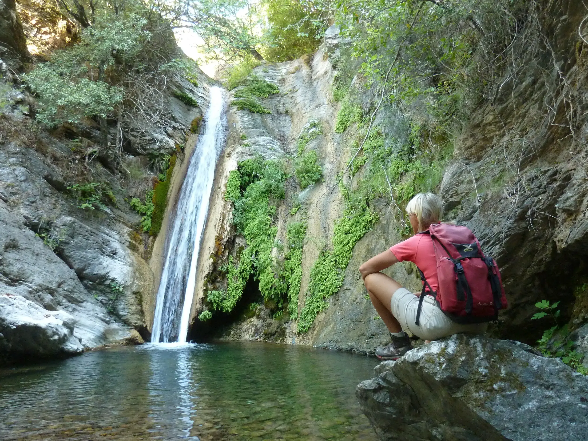 Eine Wanderin mit Rucksack macht auf einem Felsen Rast und blickt auf den Wasserfall Su Stampu de Su Turrunu in Sardinien.