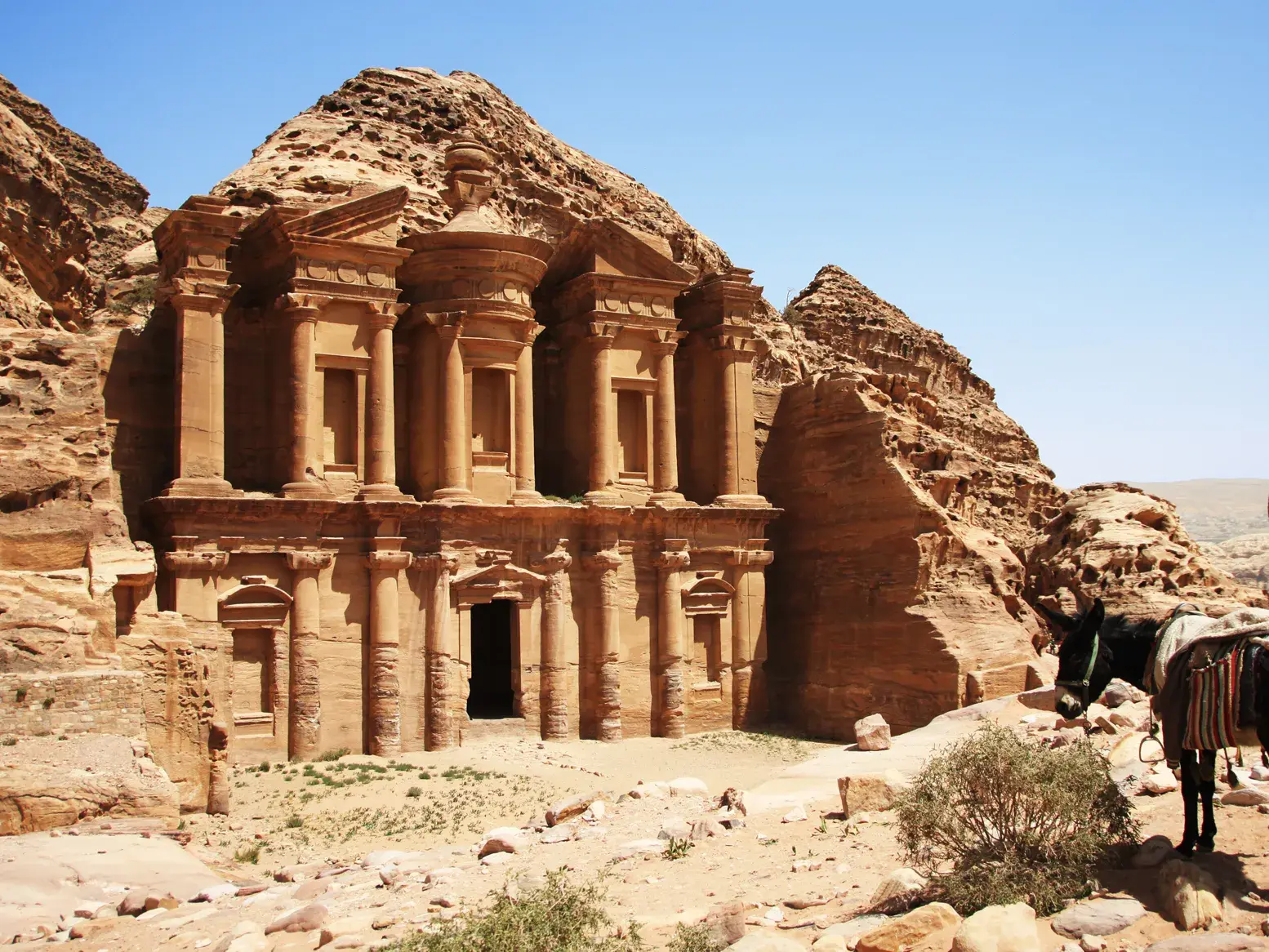 Felsentempel Ad-Deir in Petra nach der Wanderung. Ein Esel steht in der sonnigen Felslandschaft Jordaniens.