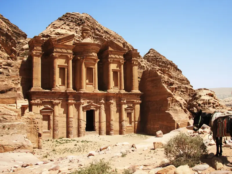 Felsentempel Ad-Deir in Petra nach der Wanderung. Ein Esel steht in der sonnigen Felslandschaft Jordaniens.