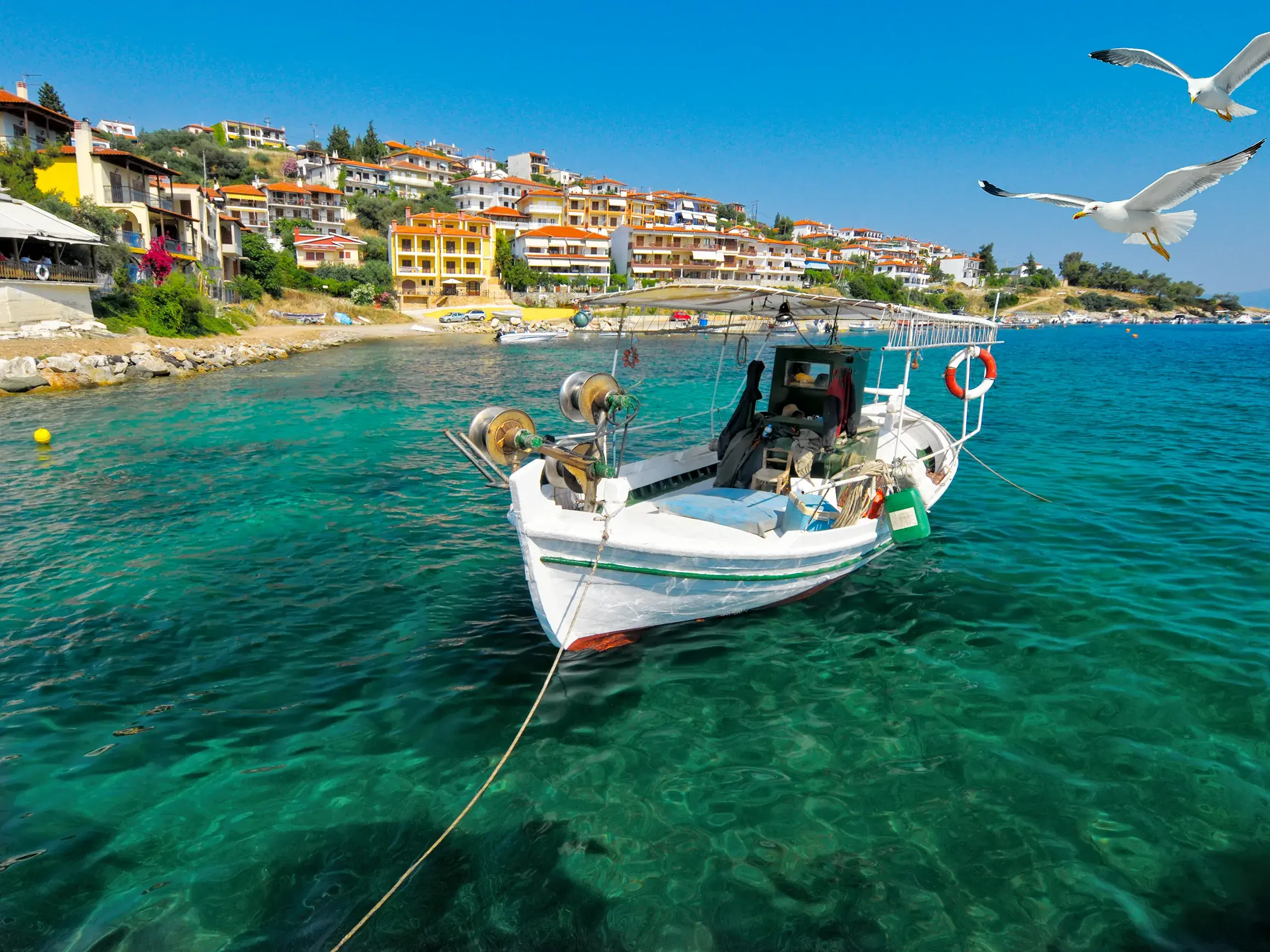Weißes Fischerboot ankert in türkisem Wasser vor einem Fischerdorf auf der Halbinsel Chalkidiki in Nordgriechenland.