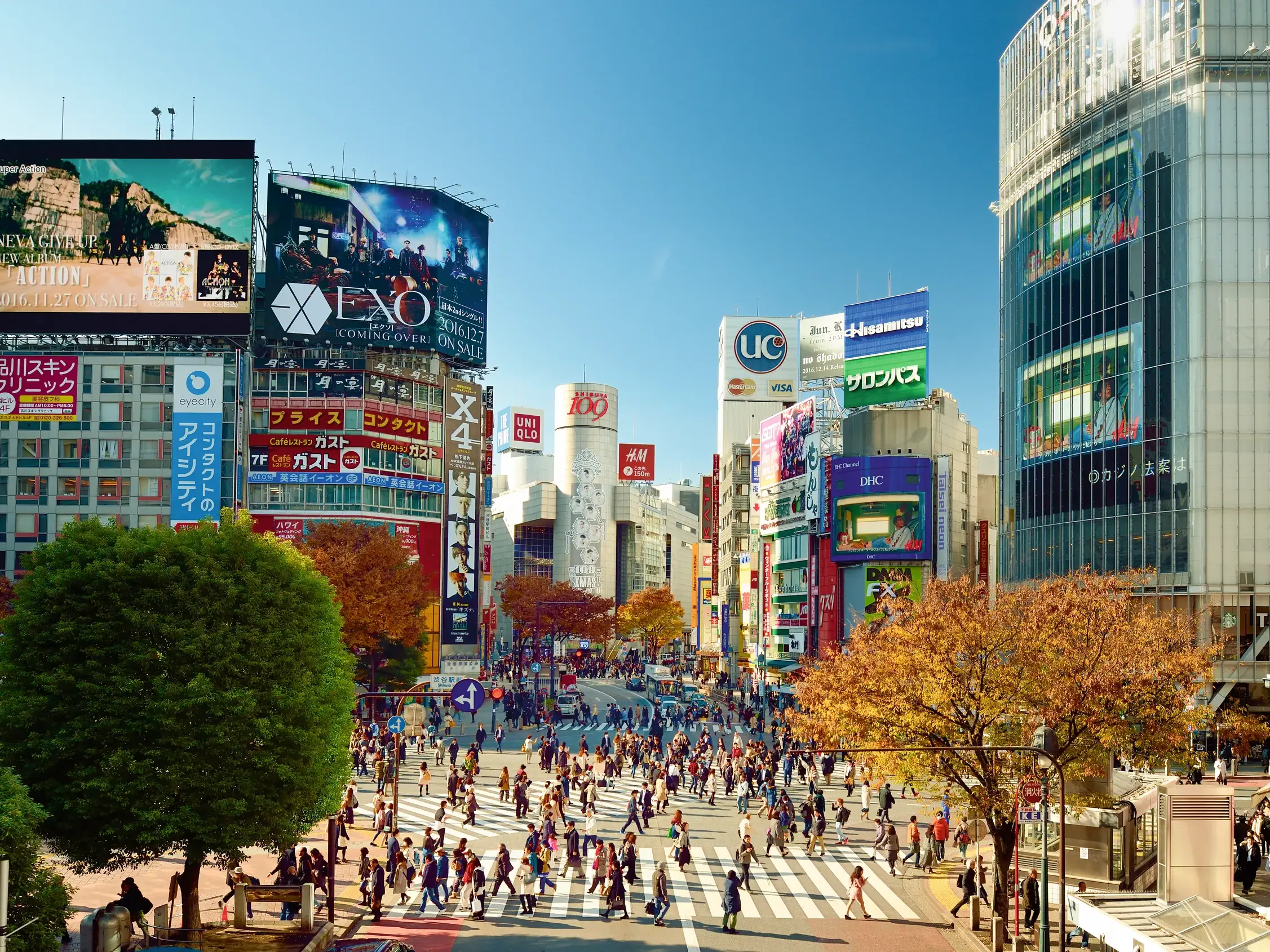 Eine Menschenmenge überquert die belebte Shibuya-Kreuzung in Tokio, umgeben von hohen Gebäuden mit bunten Leuchtreklamen.