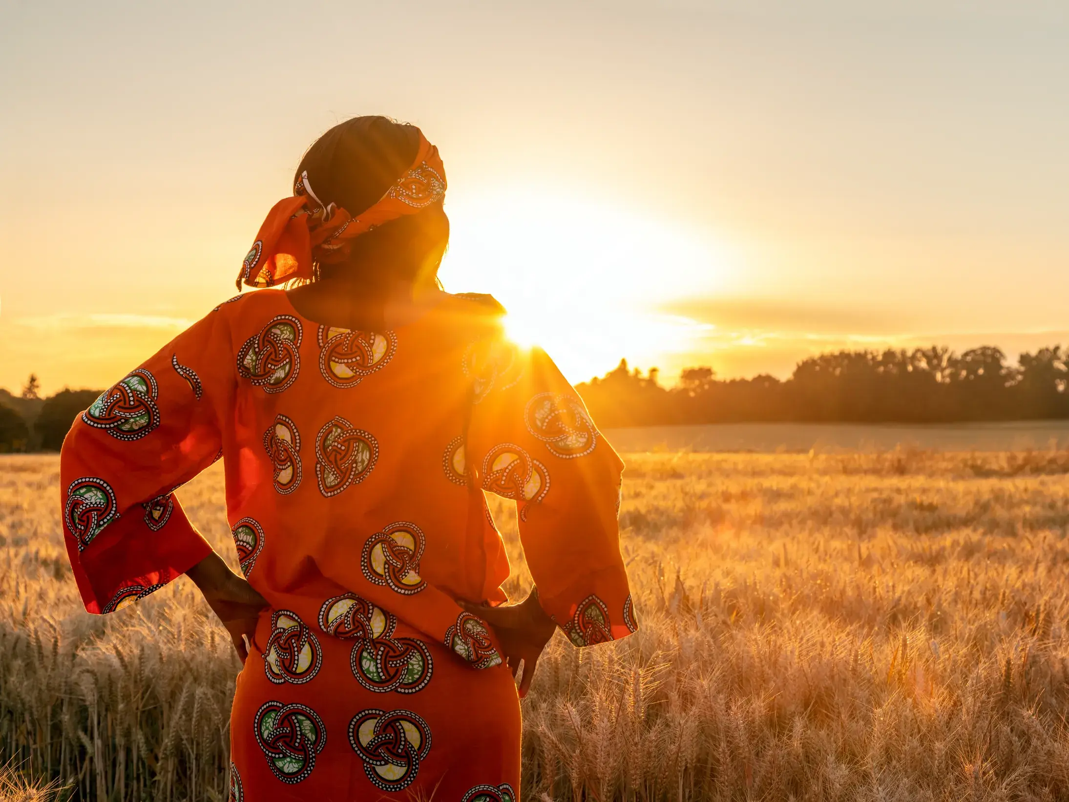 Frau in traditionellem, orangem Kleid blickt bei Sonnenuntergang über die goldenen Savannen des Hwange-Nationalparks in Simbabwe.