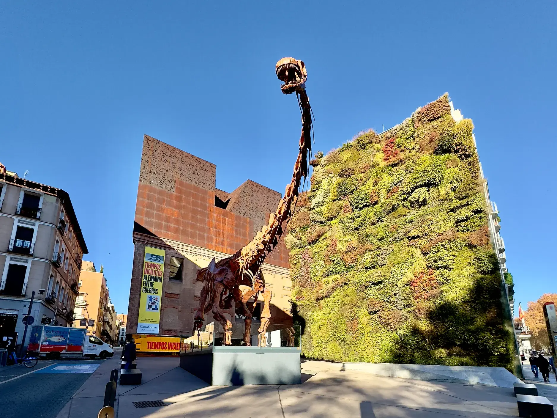 Moderne Dinosaurier-Skulptur vor der beeindruckenden, vertikal begrünten Fassade des CaixaForum in Madrid bei Sonnenschein.