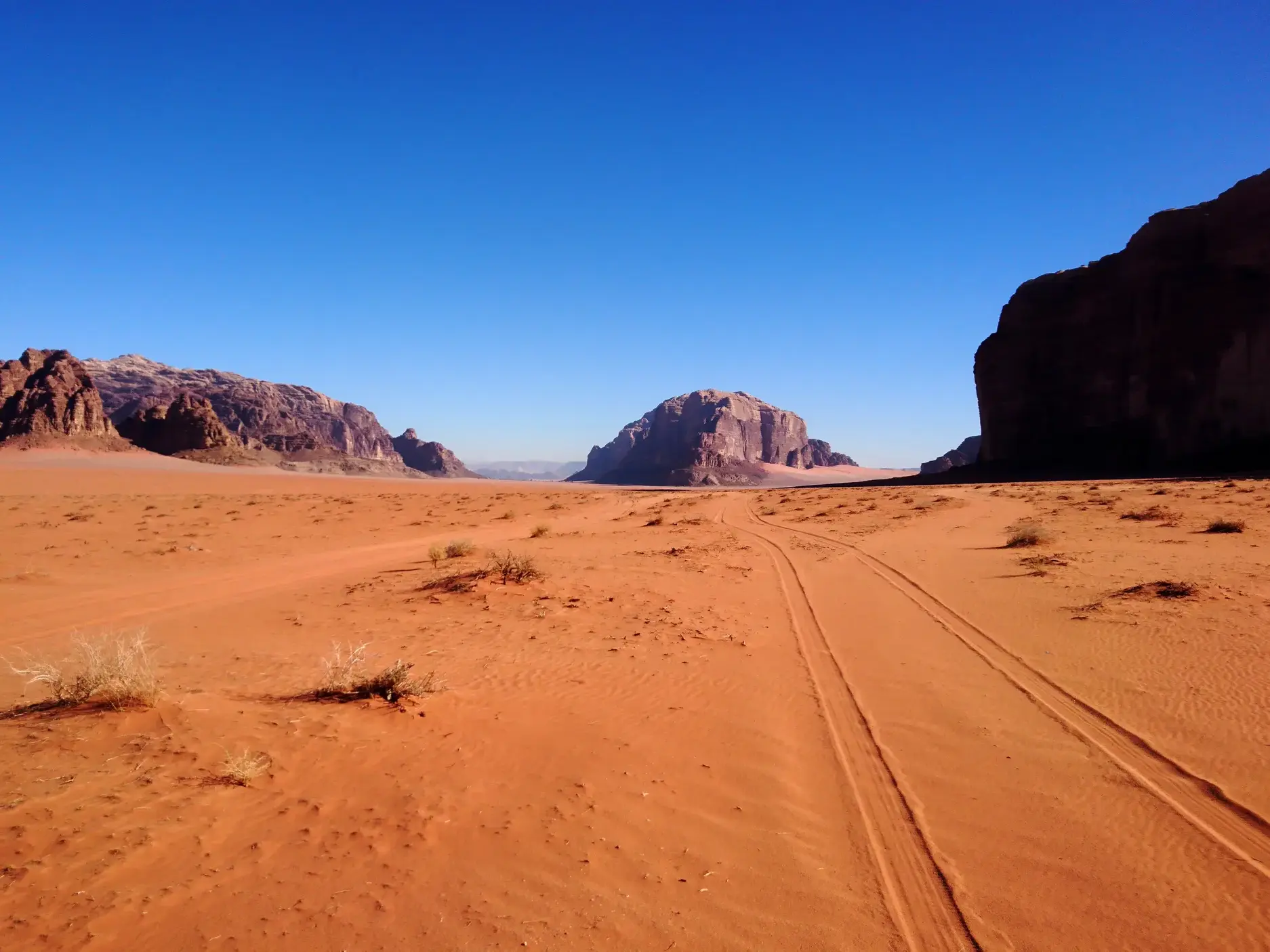 Reifenspuren führen durch den roten Sand der Wüstenlandschaft Wadi Rum zu markanten Felsformationen unter klarem blauen Himmel.