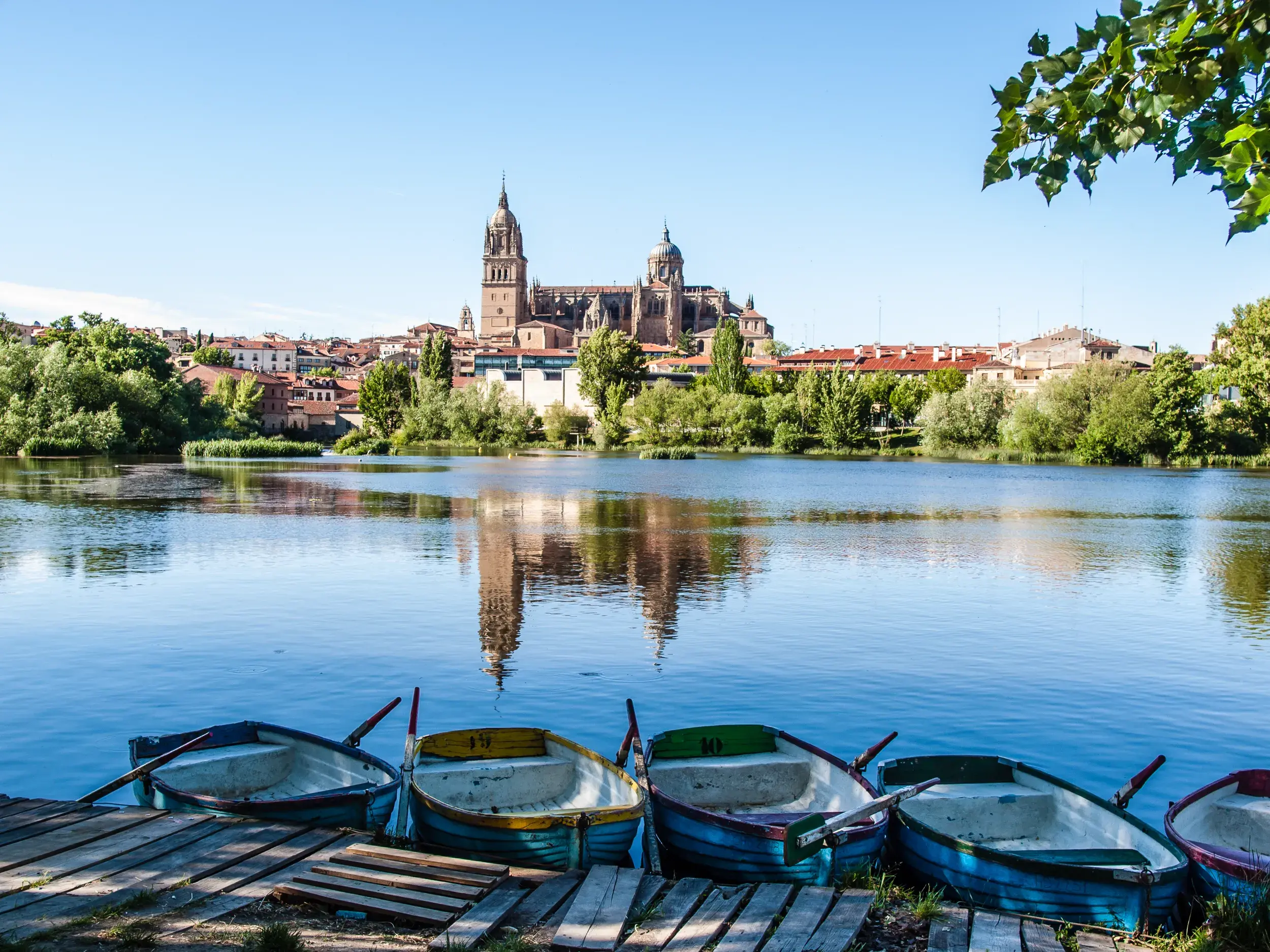 Blick über den Fluss Tormes auf die Kathedralen von Salamanca, die sich im Wasser spiegeln, mit Ruderbooten am Steg.