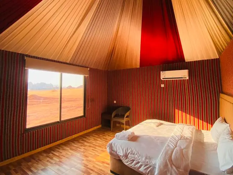 Zeltzimmer im Shaheen Luxury Camp mit bequemem Bett und großem Fenster mit Blick auf die rote Wüstenlandschaft des Wadi Ram.