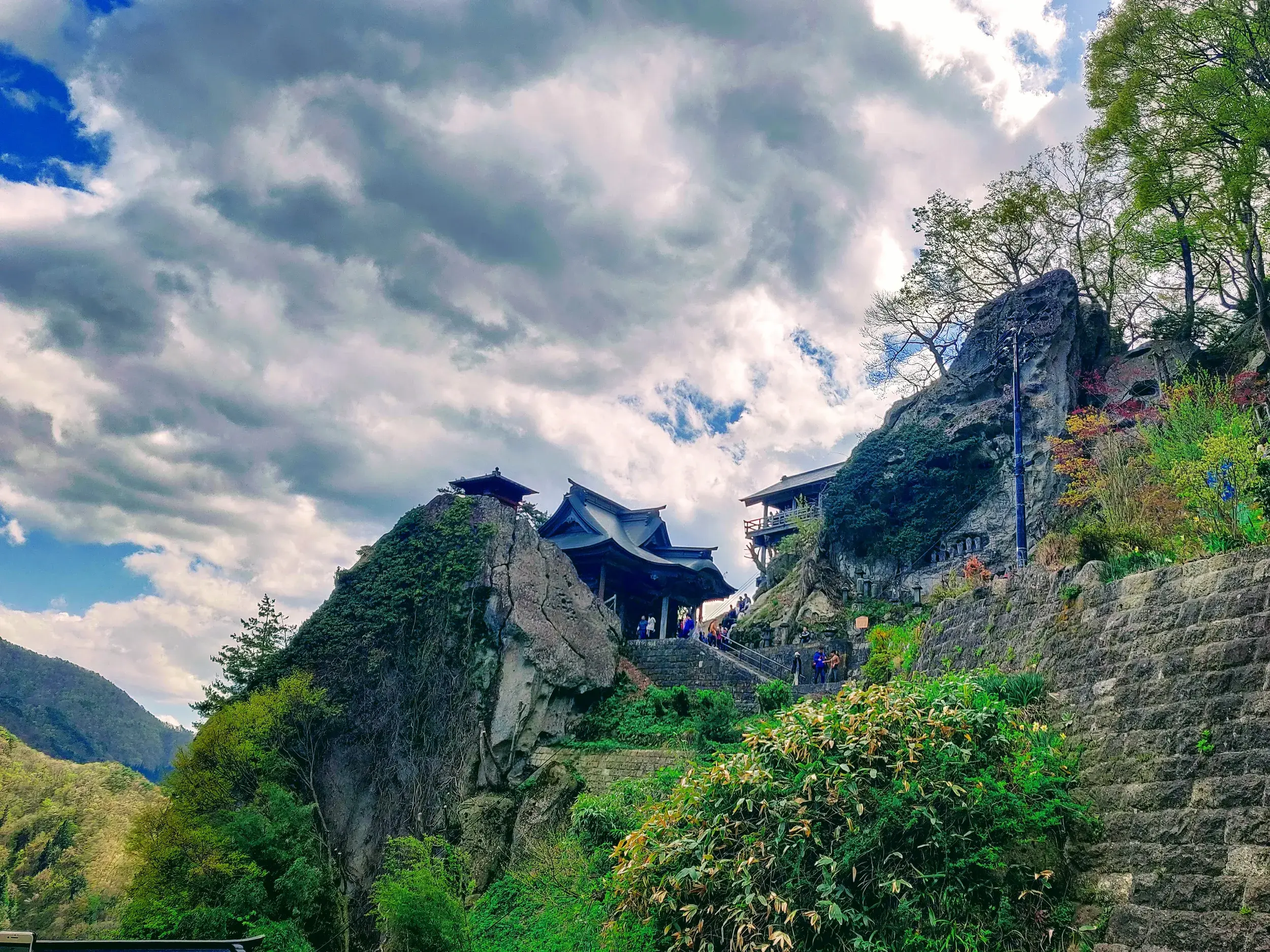 Wanderer auf dem Weg zum Yamadera Bergtempel, dessen traditionelle Gebäude sich spektakulär an eine steile Felswand schmiegen.