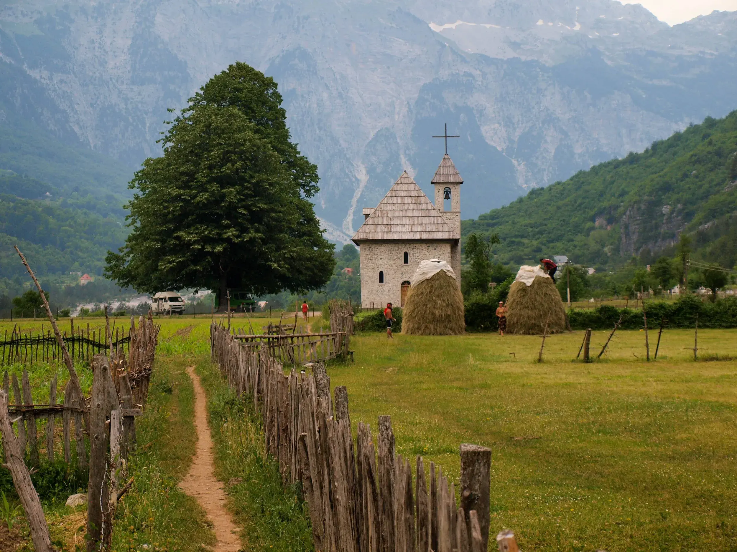 Ein Wanderweg führt vorbei an einer Steinkirche im Valbonatal, wo Menschen vor der Kulisse der Albanischen Alpen Heu ernten.
