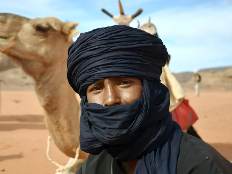 Nahaufnahme eines Tuareg-Begleiters mit blauem Chèche, im Hintergrund sein Kamel in der Wüstenlandschaft der Sahara.