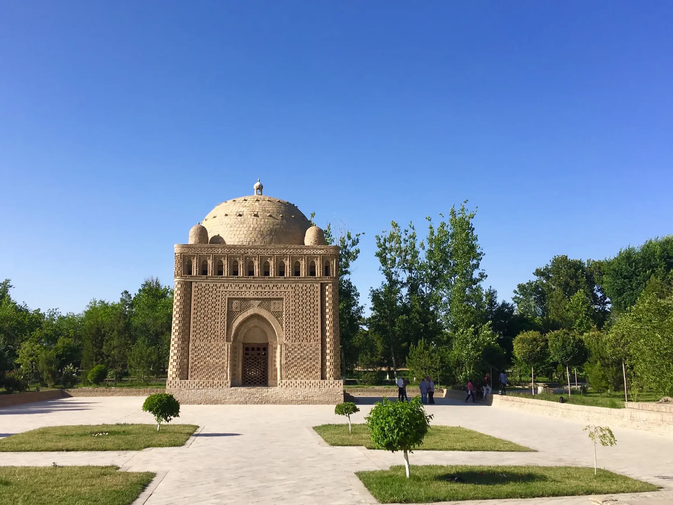 Das würfelförmige Ismail-Samani-Mausoleum in Buchara, umgeben von einem Park mit grünen Bäumen unter klarem Himmel.