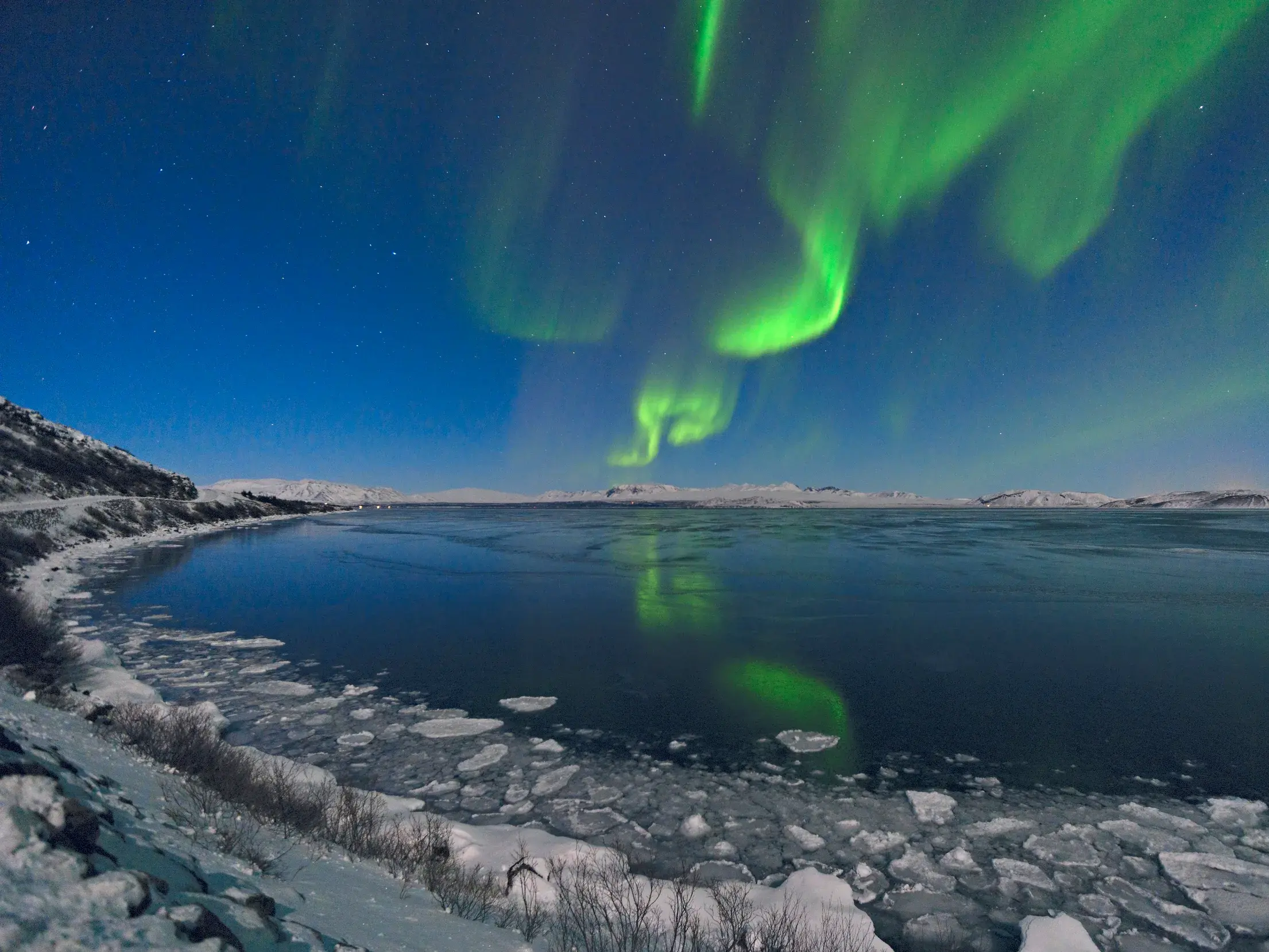 Grüne Polarlichter leuchten am Nachthimmel über schneebedeckten Bergen und eisigem Wasser in Island.