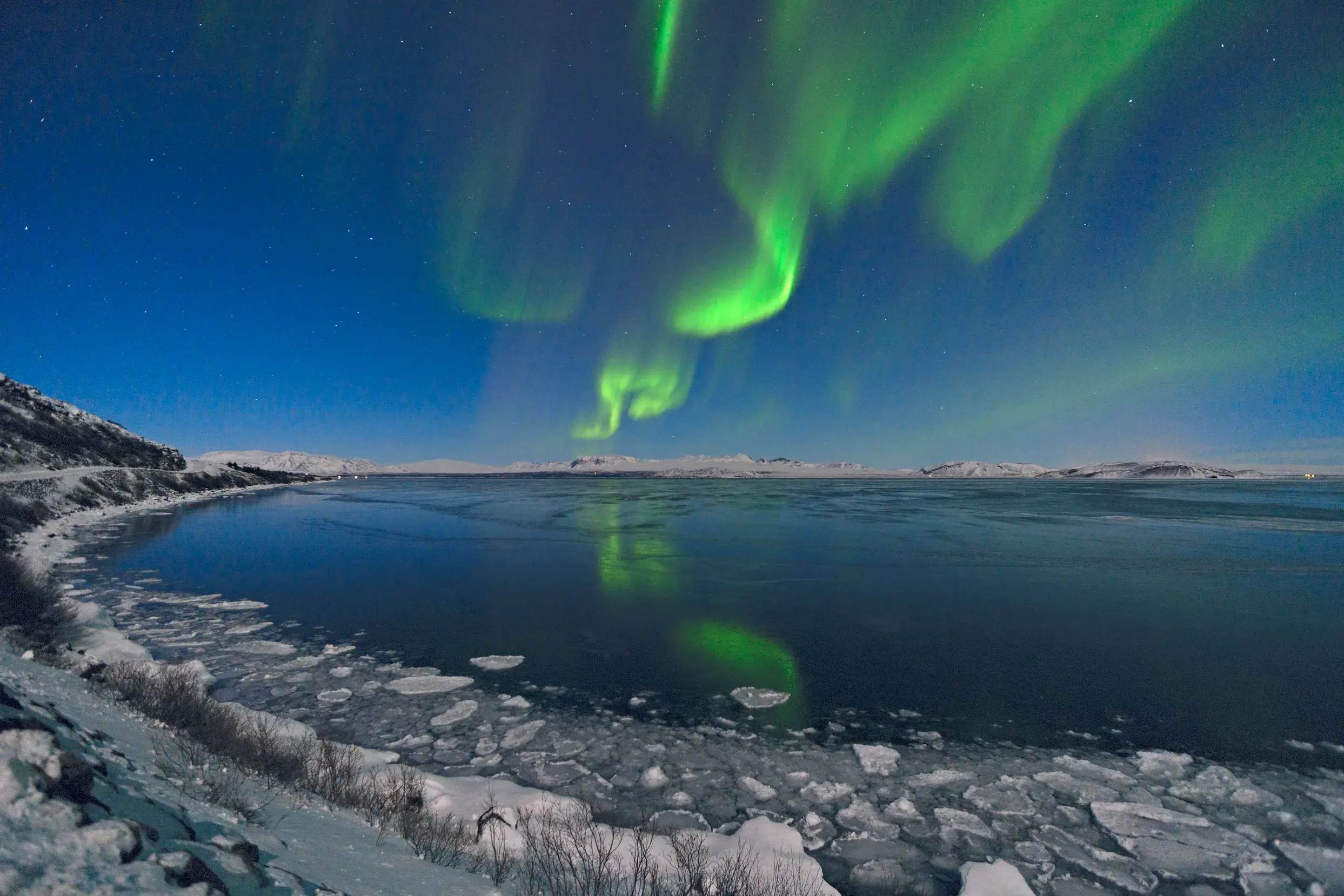 Grüne Polarlichter leuchten am Nachthimmel über schneebedeckten Bergen und eisigem Wasser in Island.