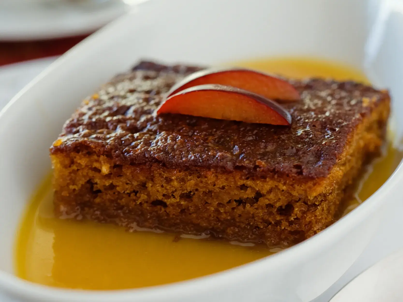 Ein Stück südafrikanischer Malva Pudding in einer weißen Schale, übergossen mit warmer Soße und garniert mit Obstspalten.