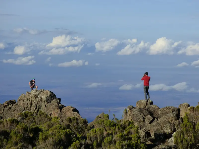 Zwei Wanderer auf felsigen Gipfeln des Northern Circuit am Kilimanjaro genießen die Aussicht über die weite Wolkenlandschaft.