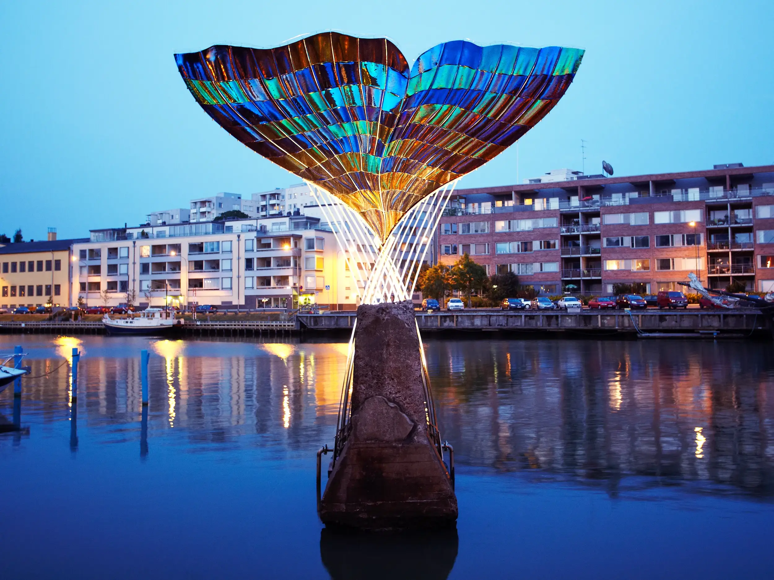 Abenddämmerung in Turku mit der bunten, beleuchteten Skulptur Harmonia, die sich im Fluss Aura spiegelt.