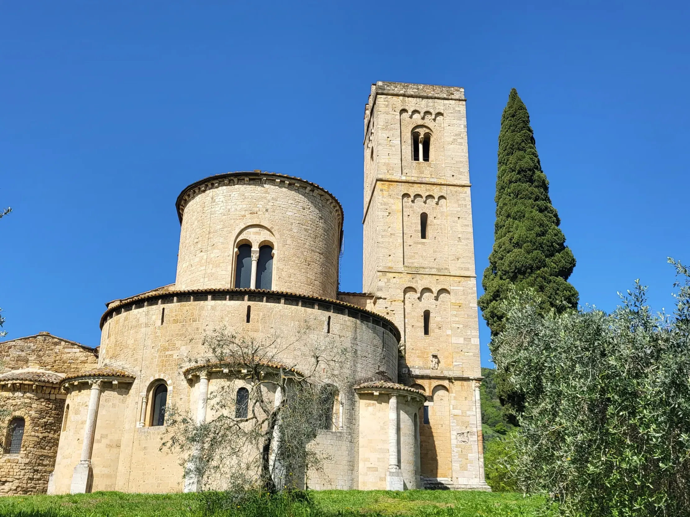 Die romanische Abtei Sant Antimo im Val d'Orcia, umgeben von Olivenbäumen und einer Zypresse unter klarem blauen Himmel.