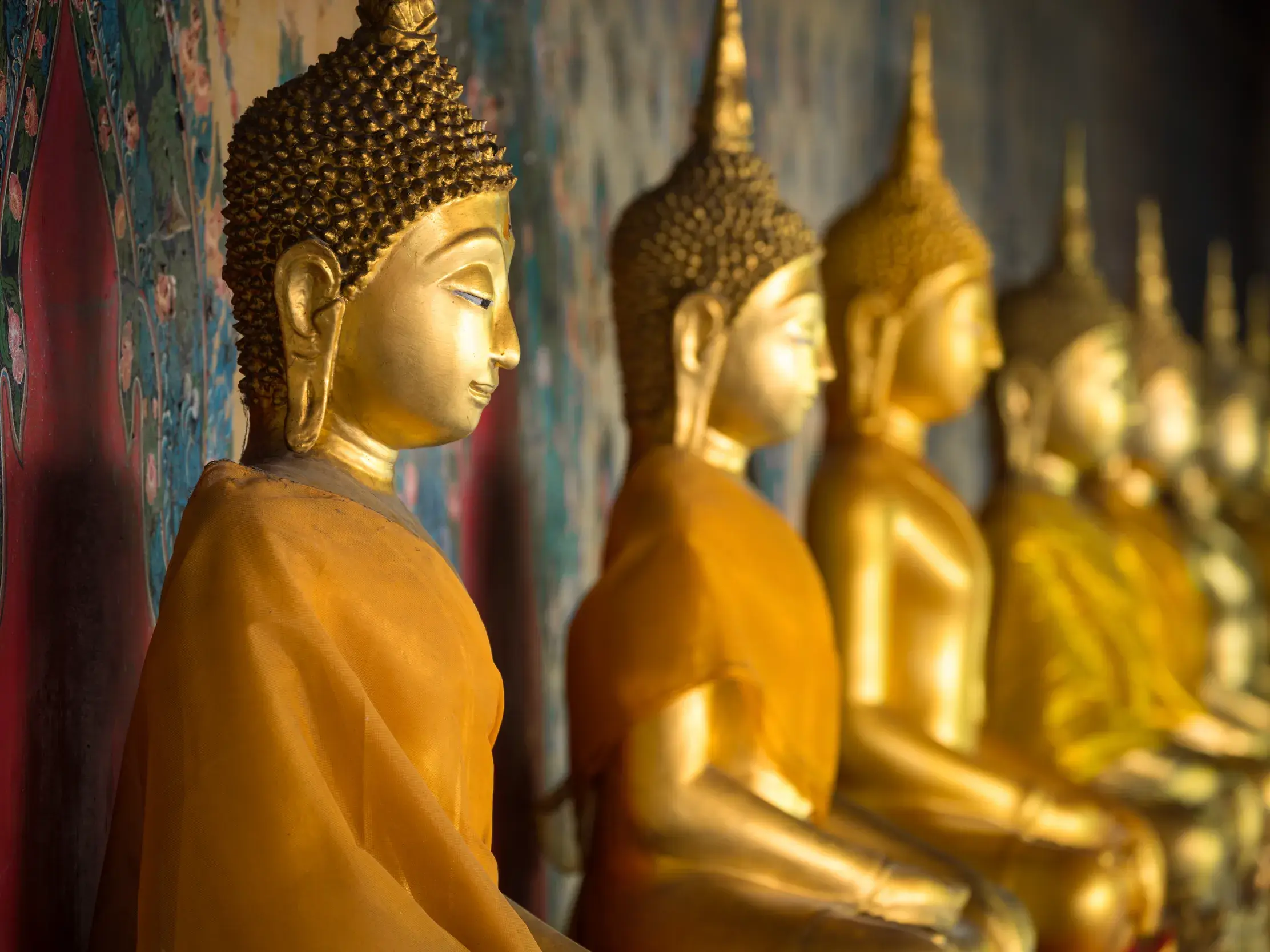 Eine Reihe goldener Buddha-Statuen mit orangefarbenen Roben sitzt in einem stimmungsvollen Tempel in Bangkok, Thailand.