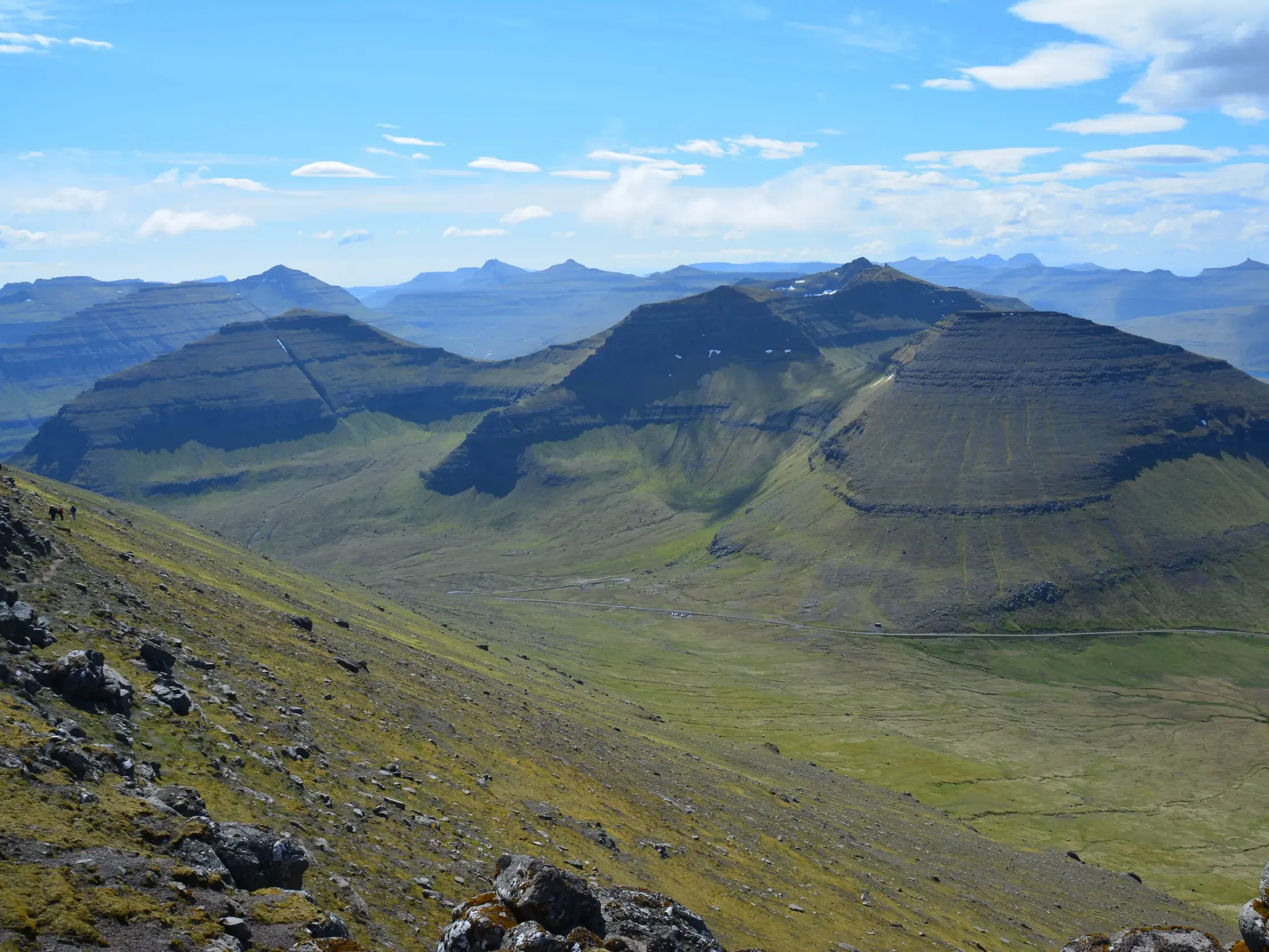 Zwei Wanderer blicken vom höchsten Berg Slættaratindur über die weite, grüne Terrassen-Berglandschaft der Färöer-Inseln.
