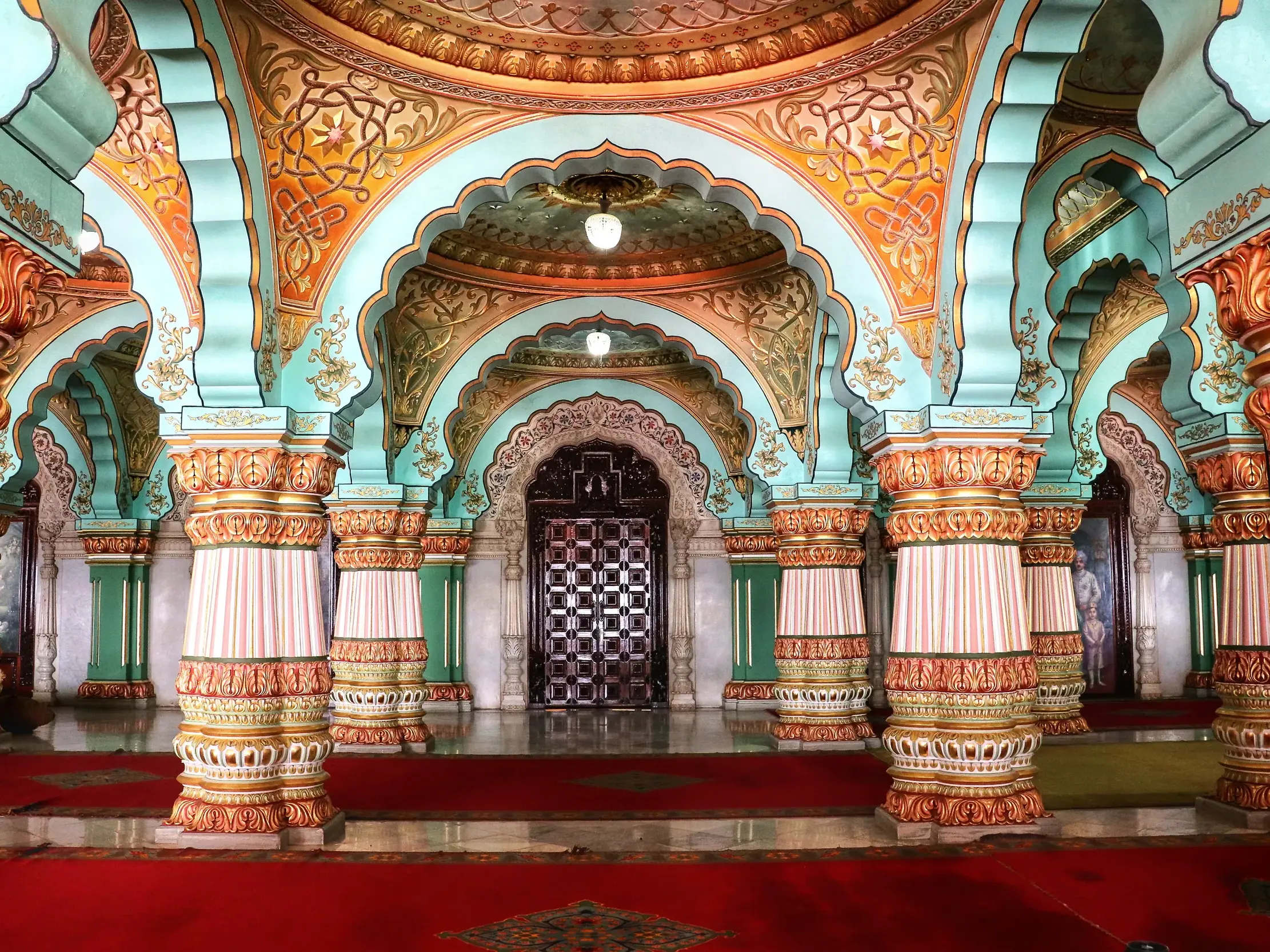 Prunkvolle Halle im Maharadschapalast von Mysore mit türkisfarbenen Bögen und kunstvoll bemalten Säulen auf rotem Teppich.