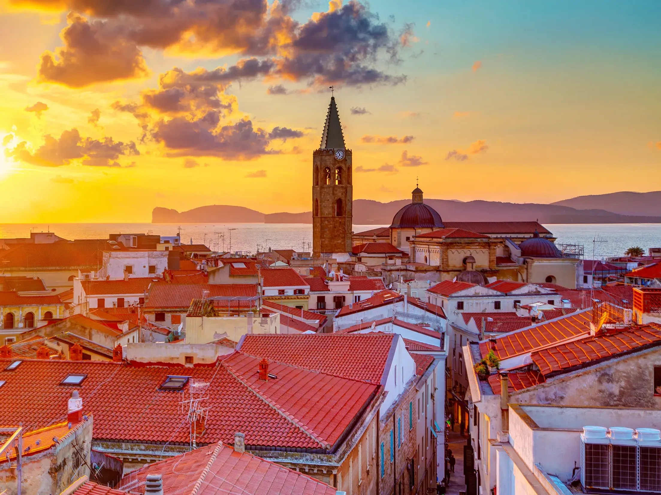 Goldener Sonnenuntergang über den roten Ziegeldächern der Altstadt von Alghero mit Blick auf die Kathedrale und das Meer.